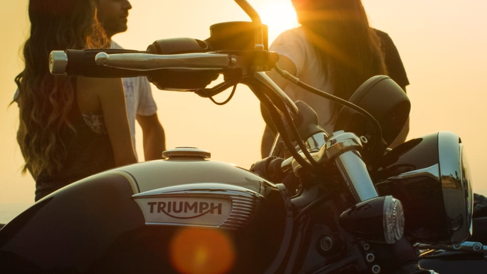 Triumph Speedmaster in the foreground showing tank and handlebars, sunset and couple in the background