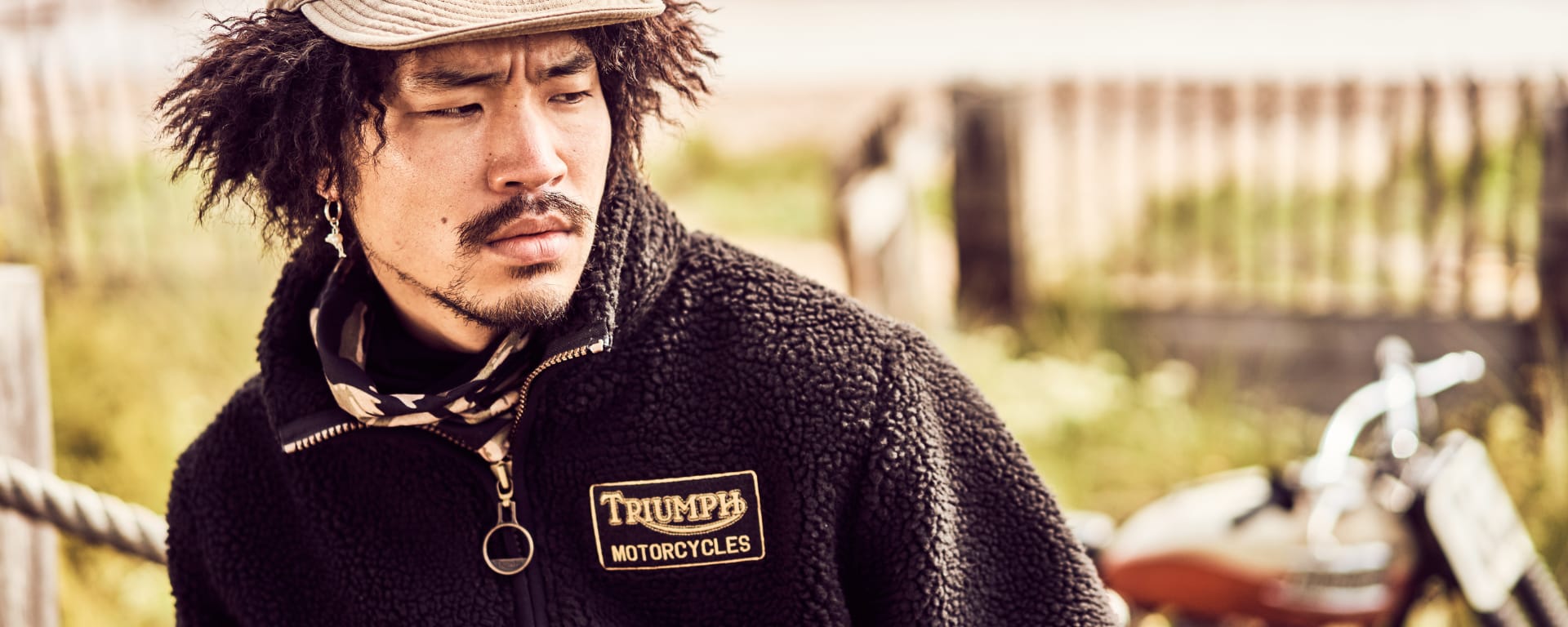 A Triumph model is wearing the Roadhouse Fleece with grassy sand dunes and a Triumph motorcycle behind them.)