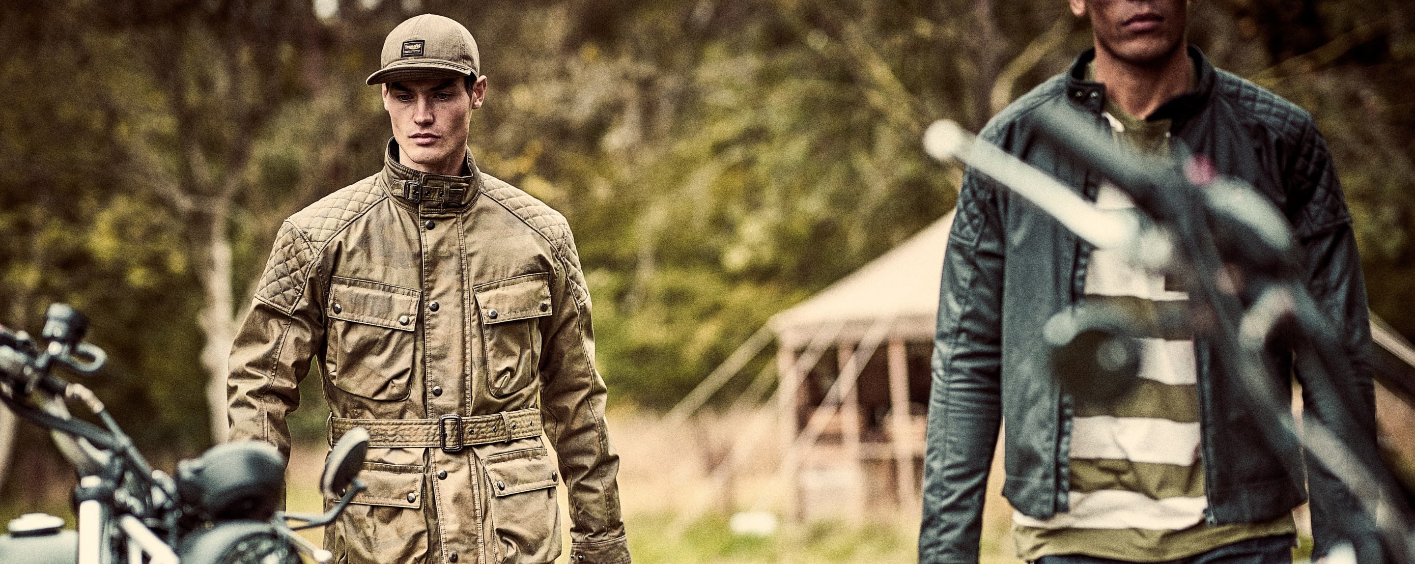 Two Triumph Motorcycle Clothing Models are walking toward motorcycles. One is wearing a Garstang Waxed Jacket in Camo, the other is wearing a Kirk Waxed Cotton Biker Jacket.)