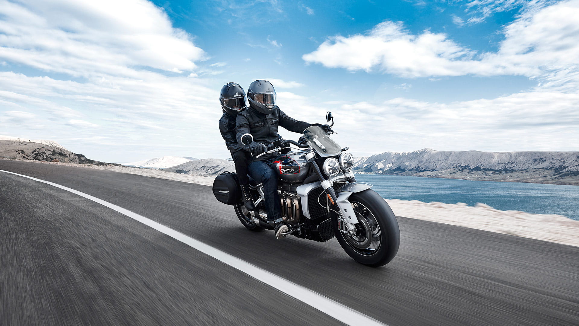 Triumph Rocket 3 GT in aluminium silver and storm grey, fitted with accessories including; black sports pannier with Triumph badging and screen, travelling along a straight road