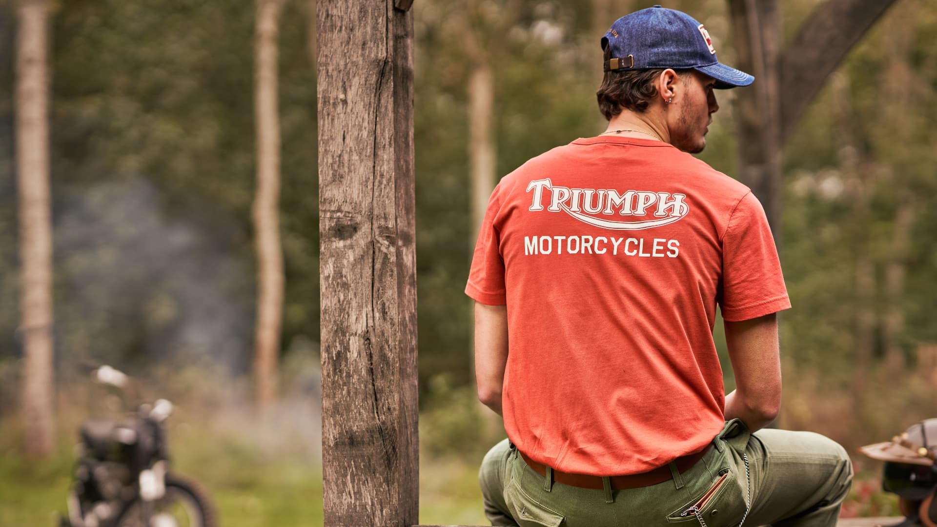 A male model is sat in the forest wearing a red Adcote t-shirt.