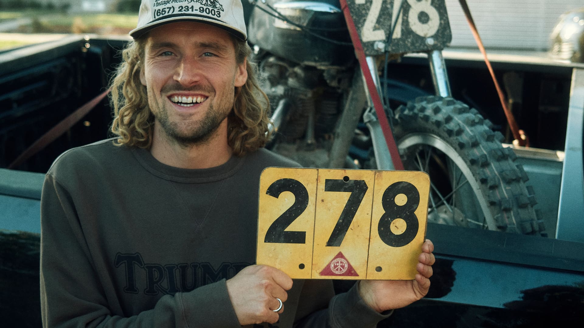 Man with racing number plate with Triumph bike in the back