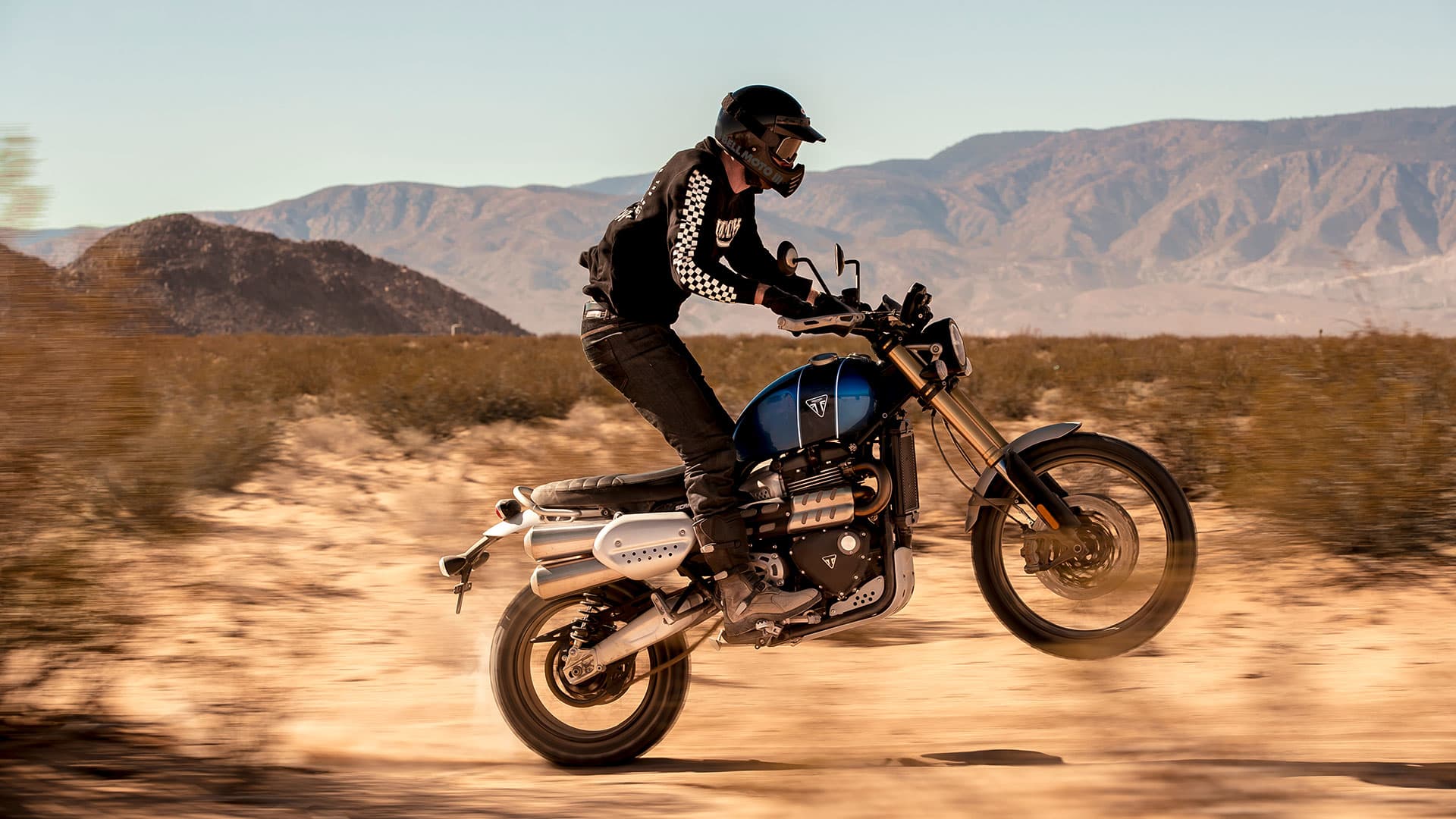 Triumph Scrambler 1200 XE scrambling across sandy plains performing a wheelie. Rider wearing limited edition Scrambler 1200 clothing