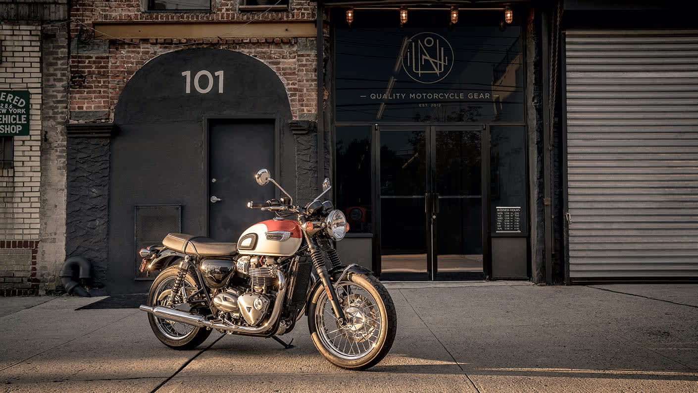 Image of Triumph Bonneville T100 in orange and white colour variant, parked in urban setting. 