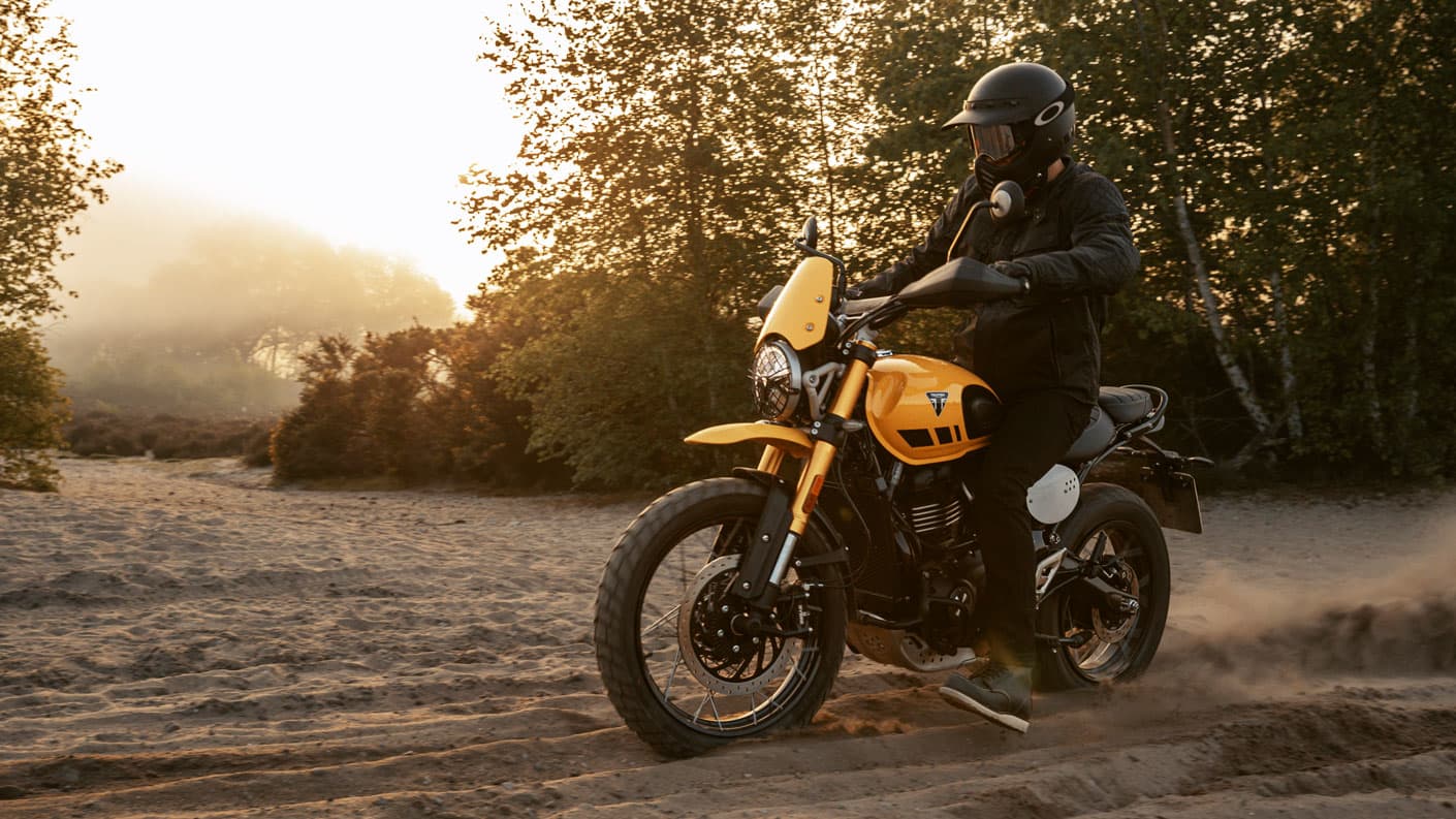 Motorcyclist rides a Racing Yellow Triumph Scrambler 400 XC over sandy terrain