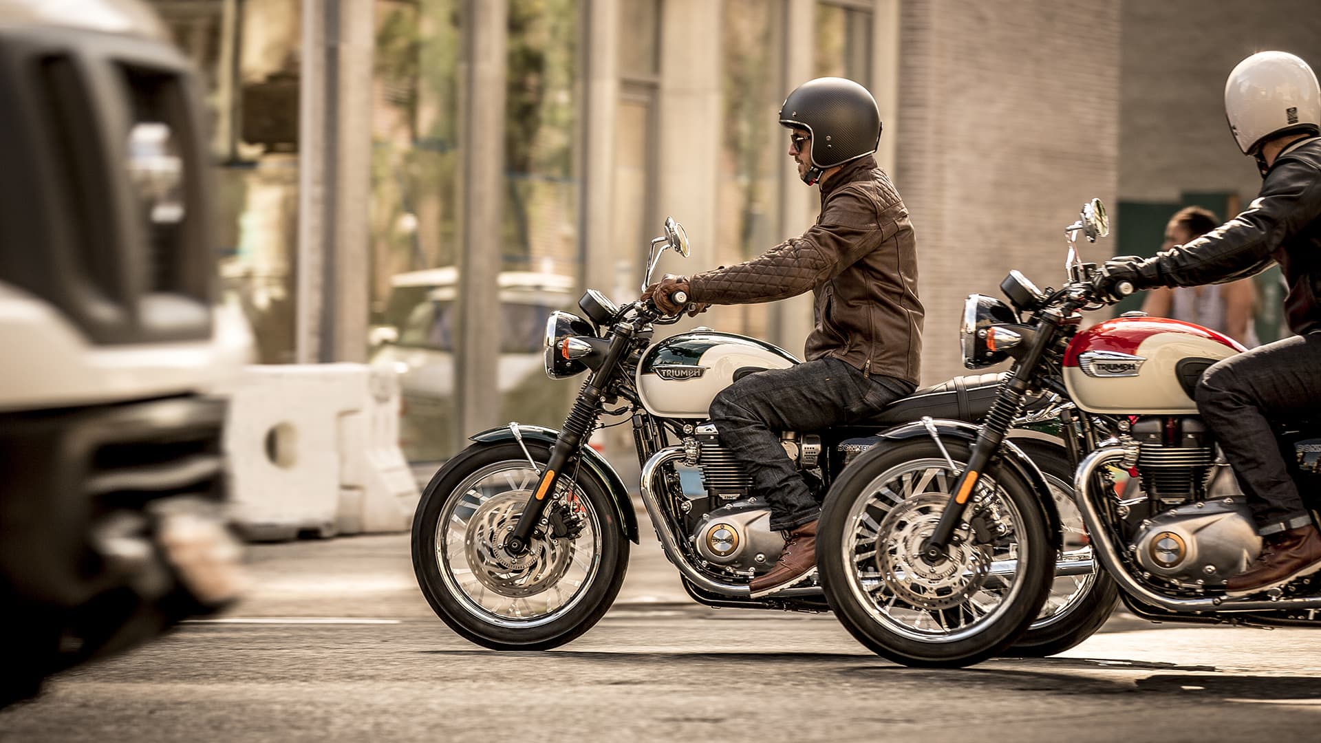 Two riders on Triumph T120s riding through city street