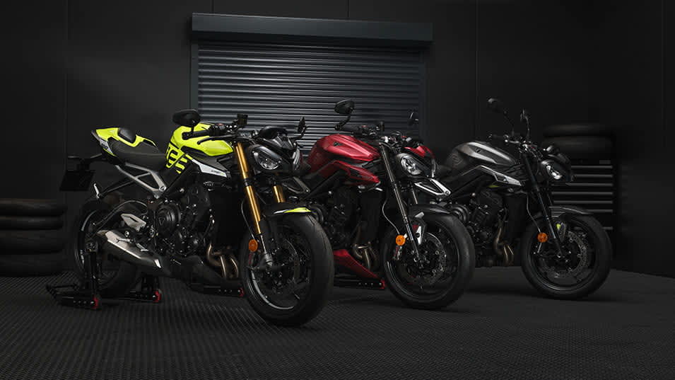 Three modern Triumph Street Triple roadster motorcycles—one in vibrant yellow, one in deep red, and one in sleek grey—parked side-by-side inside a dark, professional garage setting with a black roller door in the background.