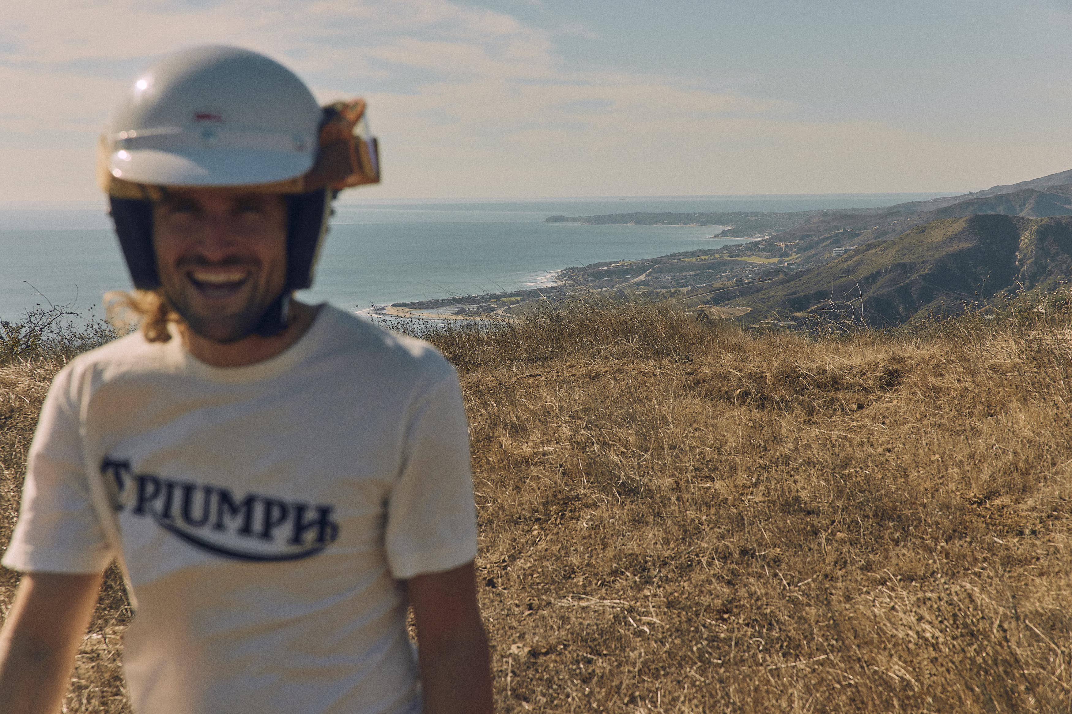 OPERATING HOURS Man in the desert smiling and wearing Triumph Motorcycles Heritage Fork Seal T-Shirt