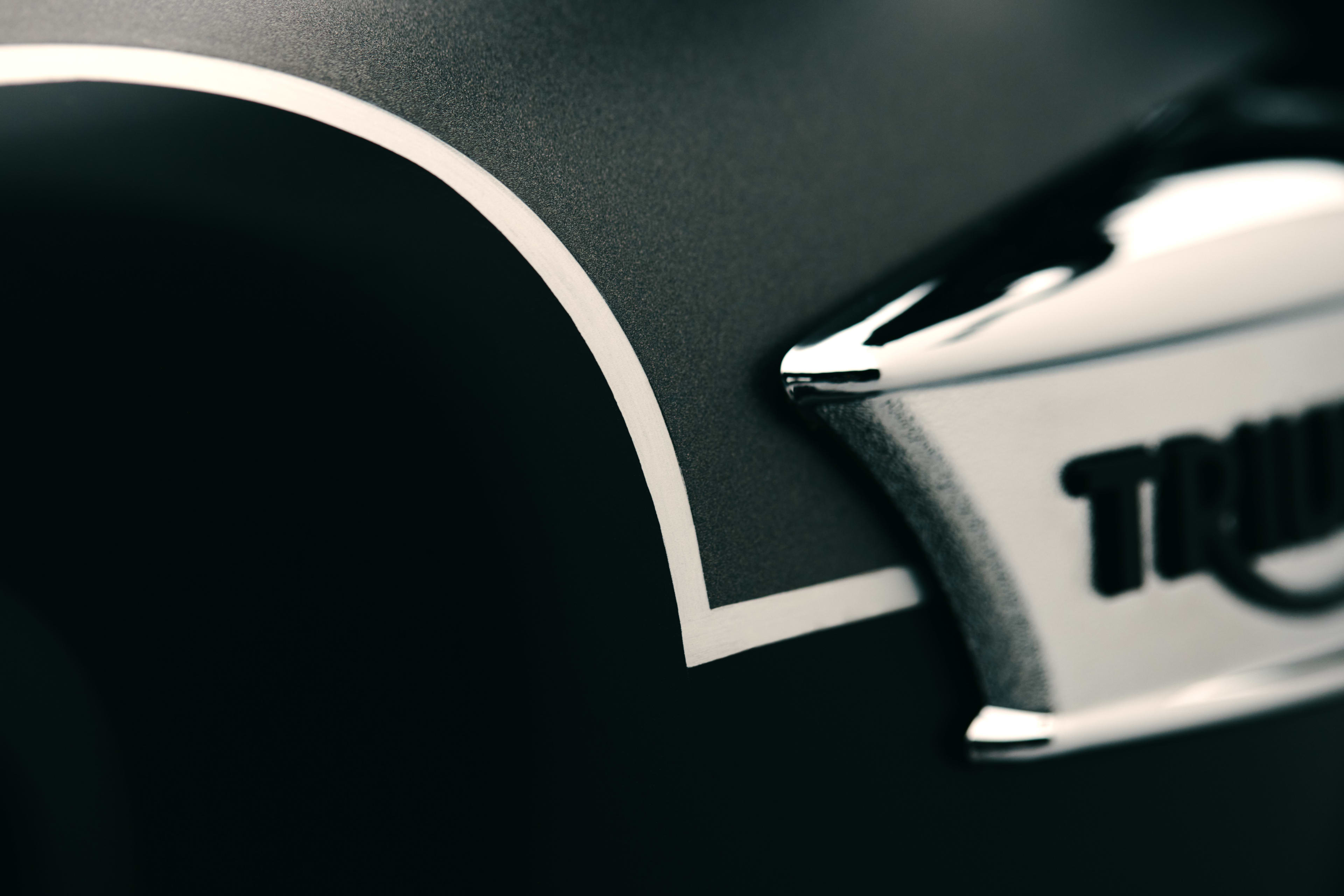 Macro detail of hand-painted coach lining separating satin grey and black paint on a Triumph fuel tank beside chrome badge.
