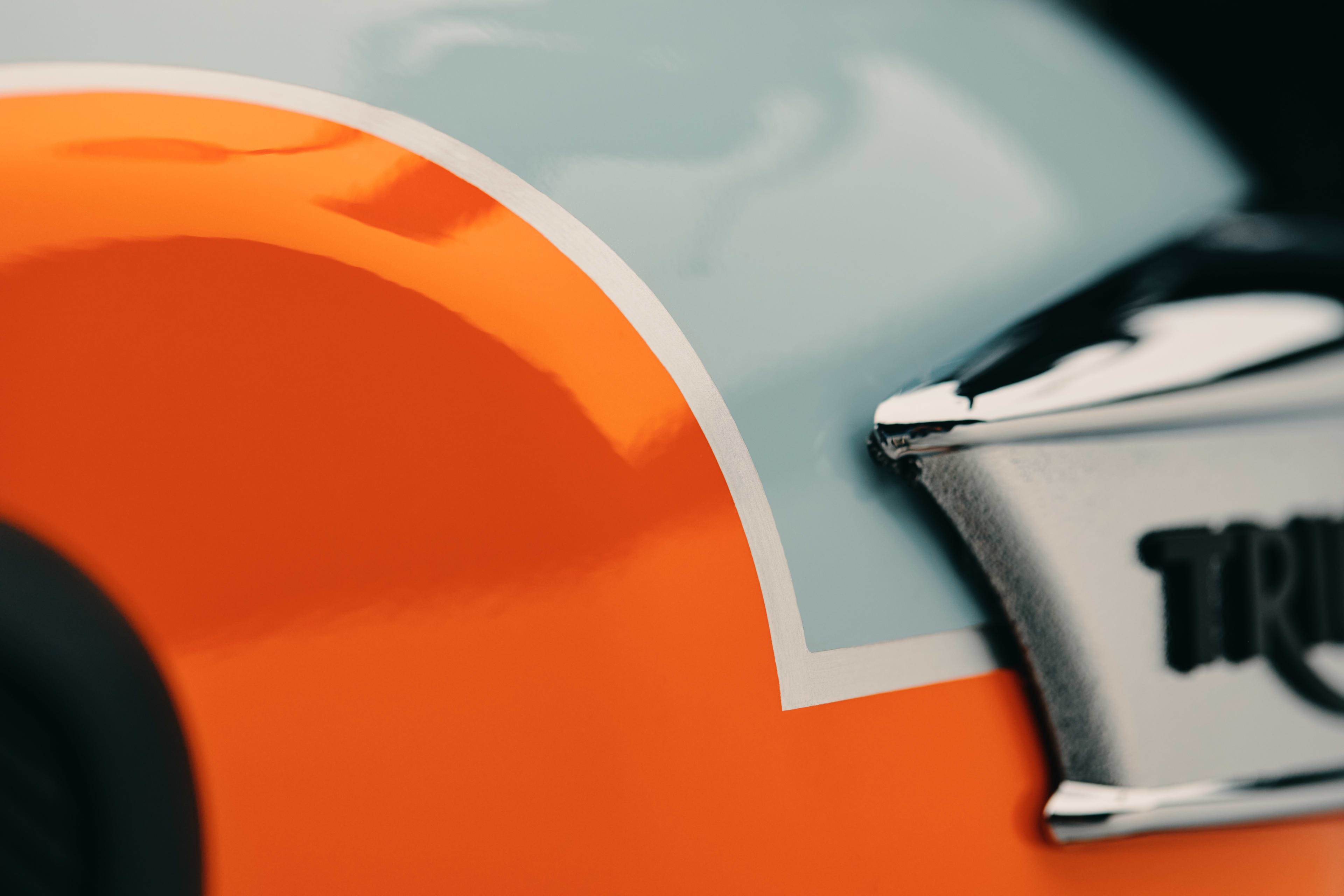Macro detail of hand-painted coach lining separating orange and light blue paint on a Triumph fuel tank beside chrome badge.