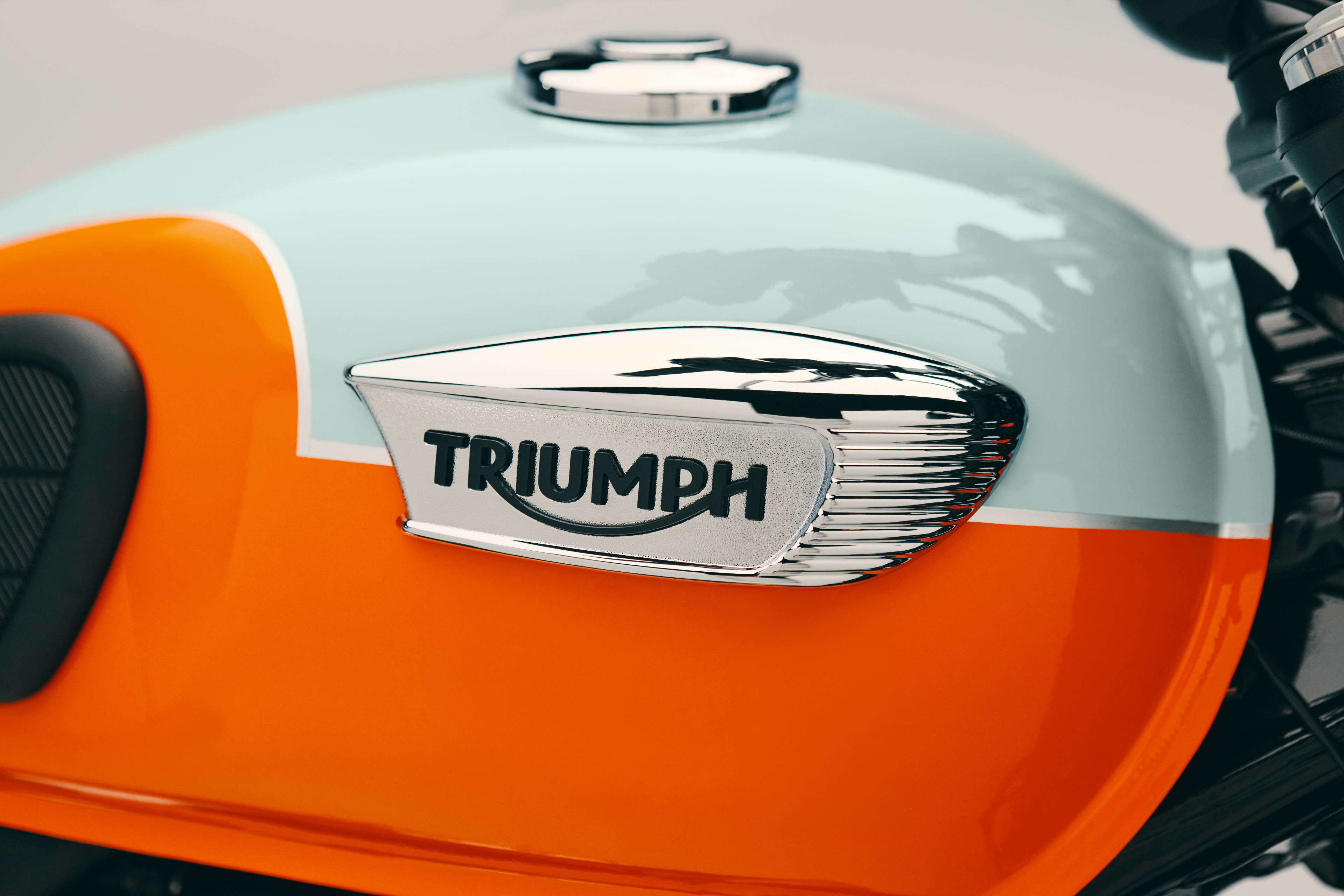 Detailed shot of a classic chrome Triumph badge on a two-tone light blue and bright orange Triumph Bonneville T40 motorcycle fuel tank.
