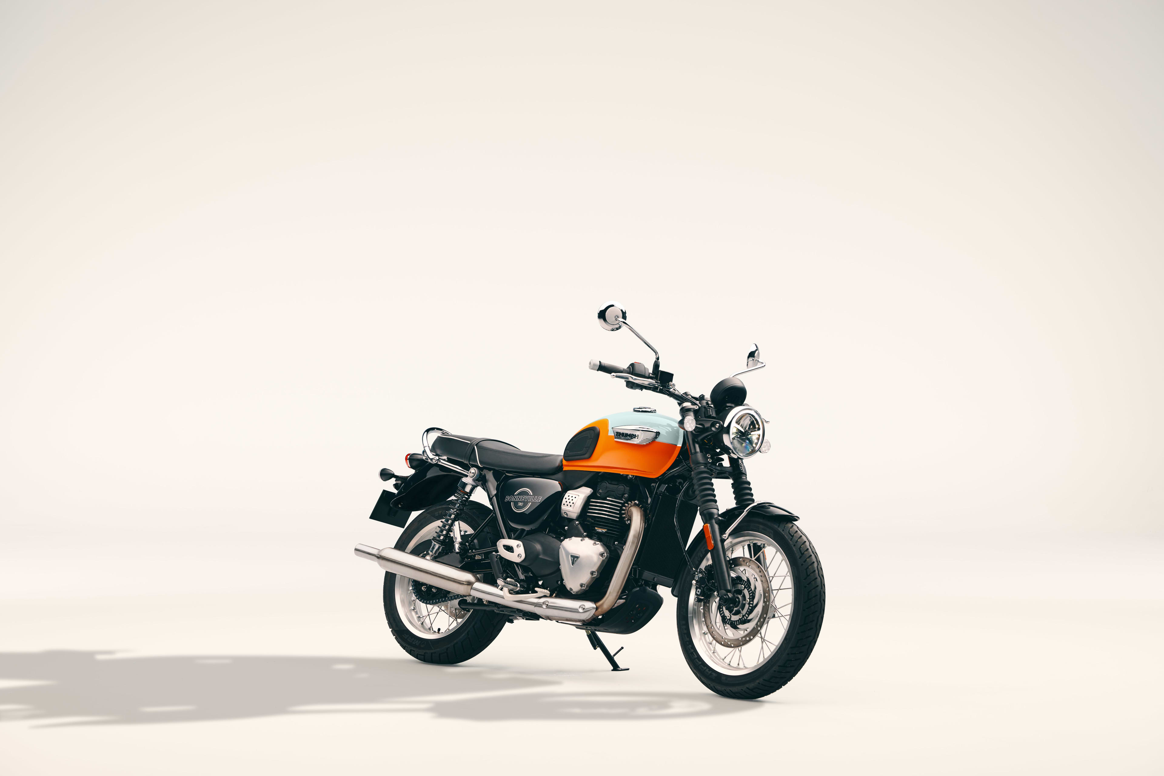 Studio shot of an orange and white Triumph Bonneville T40 motorcycle with chrome exhaust parked on a light background.