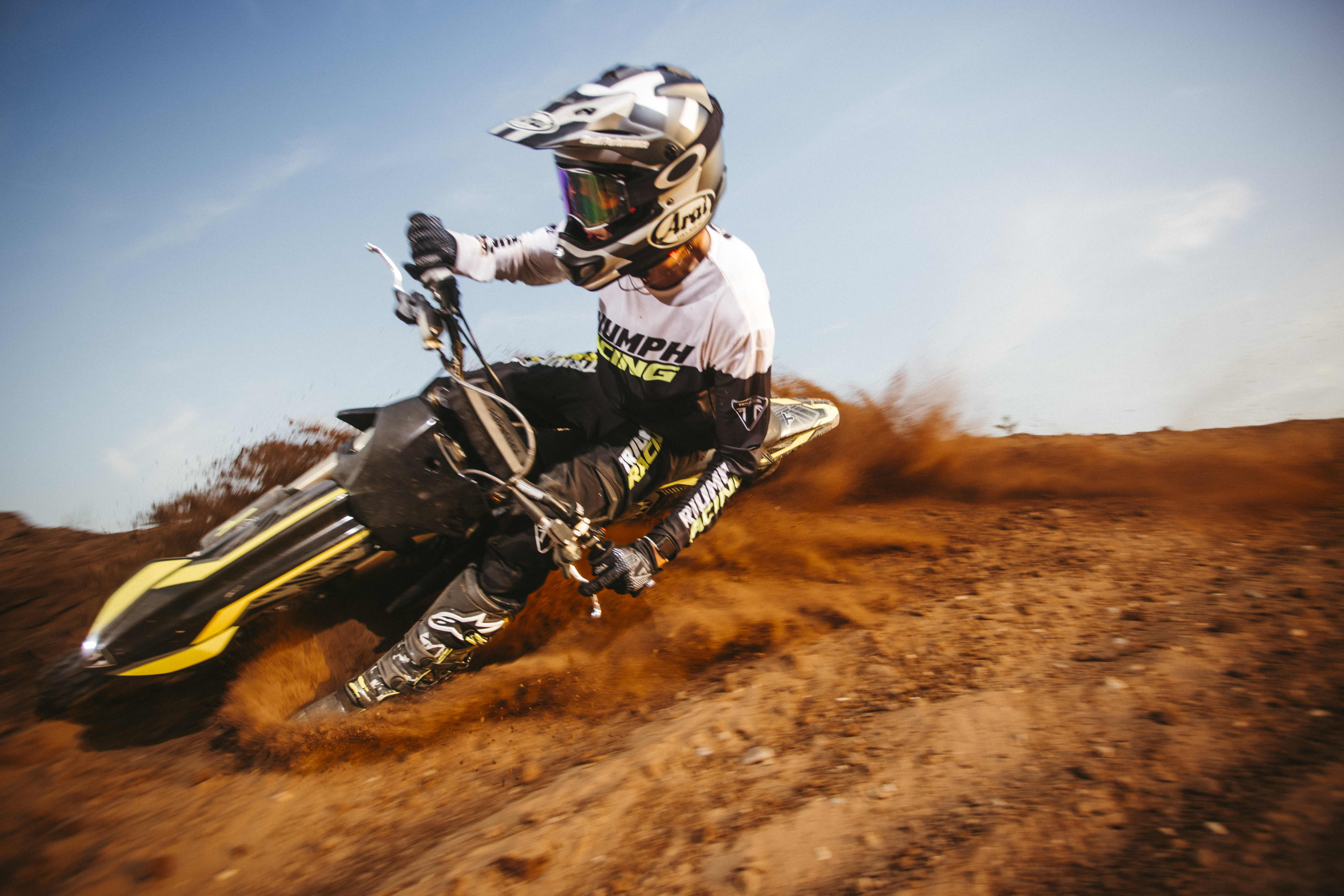 Front view of a motocross rider in full gear leaning heavily into a dirt corner, spraying up a cloud of loose soil under a clear sky.