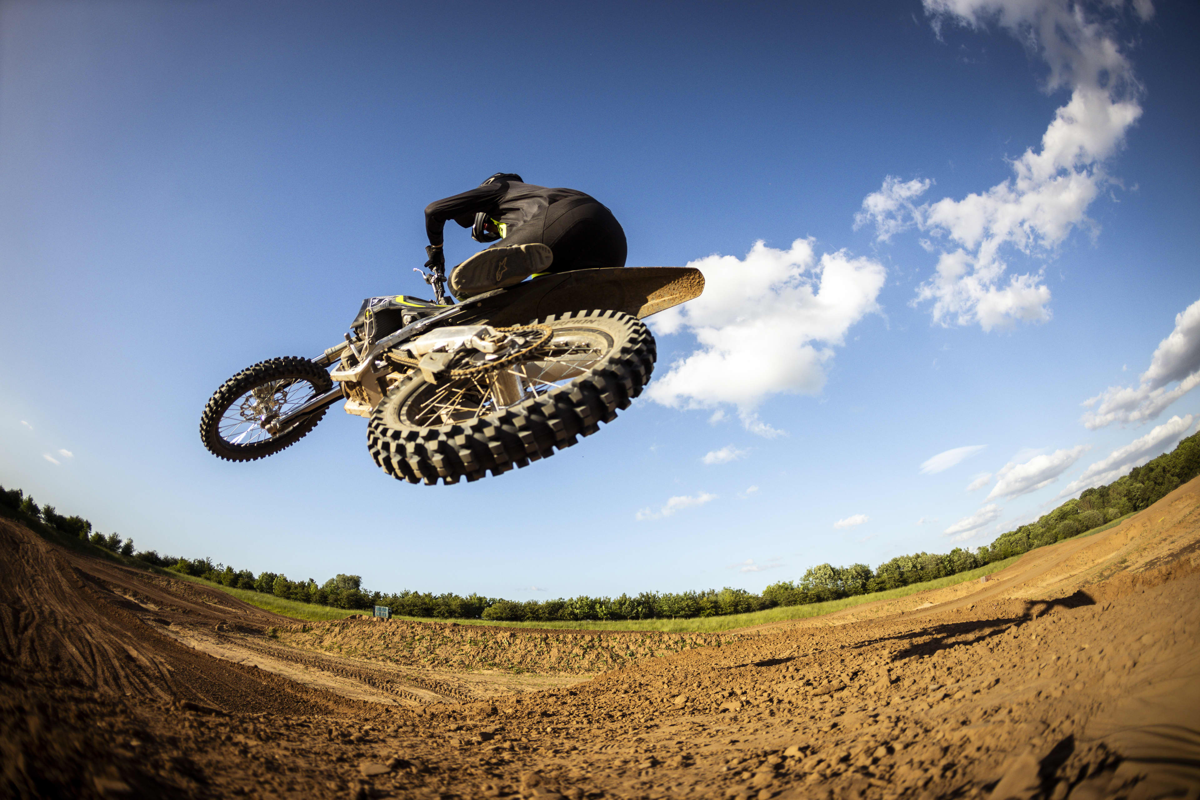 Low-angle wide fisheye perspective looking up at a rider airborne on a Triumph TF 250-X dirt bike set against a sunny sky with scattered clouds.