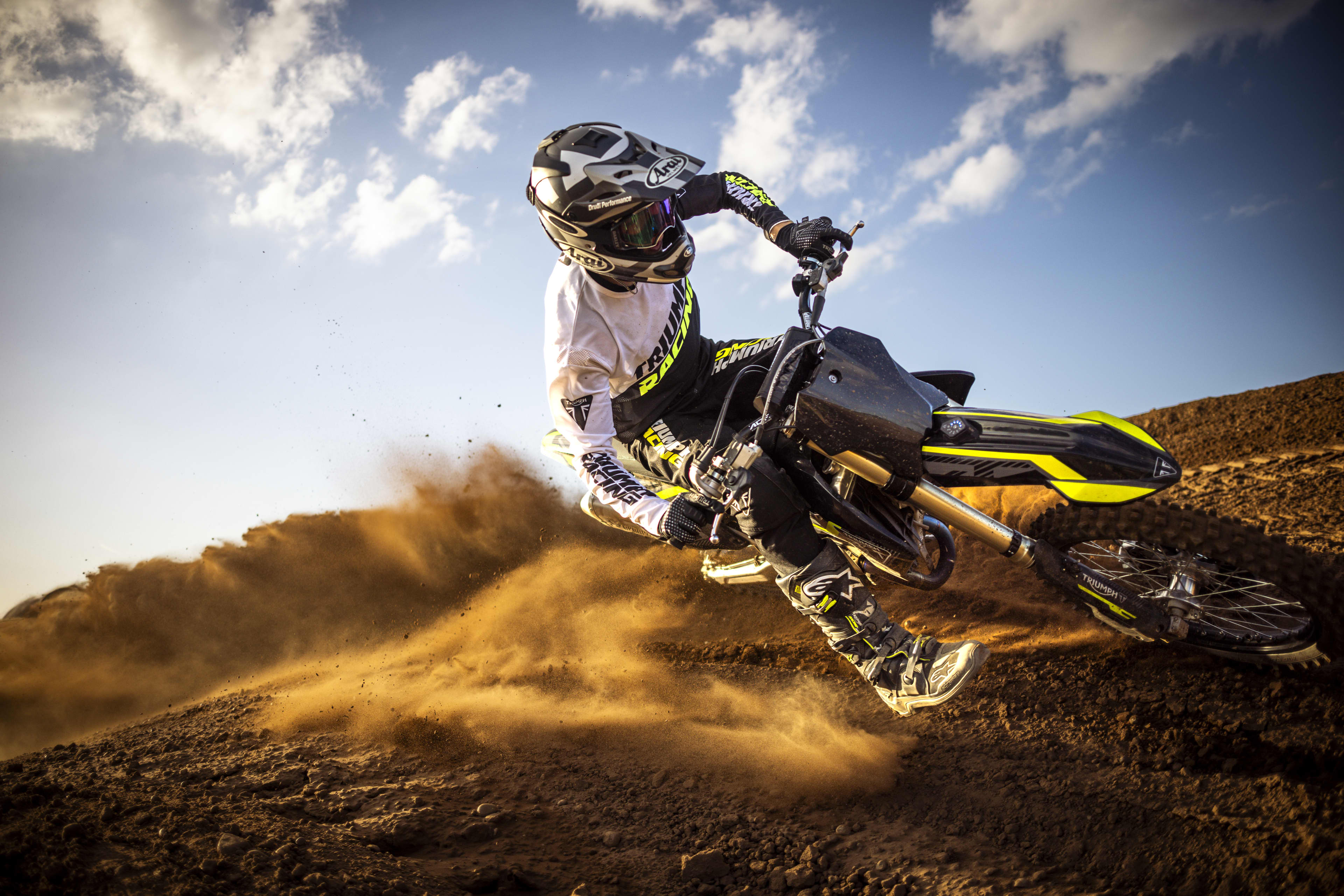 Dynamic wide action shot of a rider wearing Triumph Racing gear carving hard through a banked dirt turn on a Triumph TF 250-X motocross bike, sending up a massive cloud of dust beneath a bright sky.