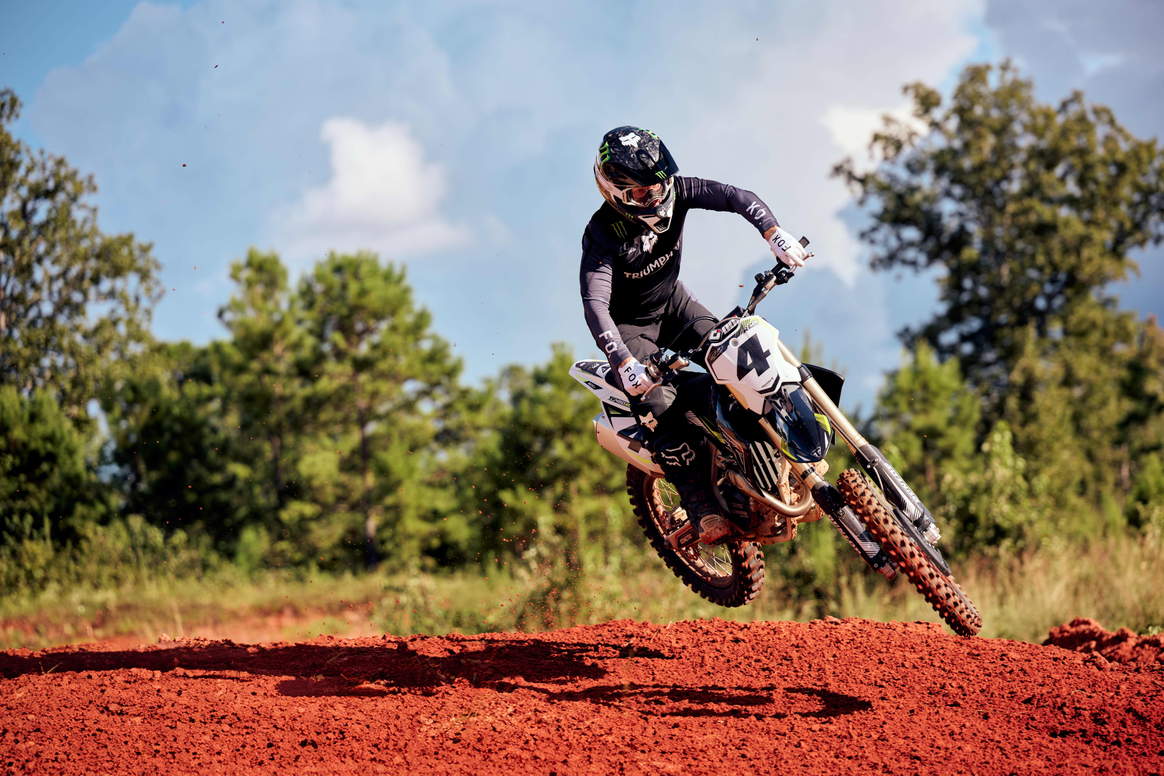 Fabricada para dominar, desarrollada para ganar Action shot of a rider in black gear catching air on a Triumph TF 450 RC dirt bike over a red dirt motocross track.