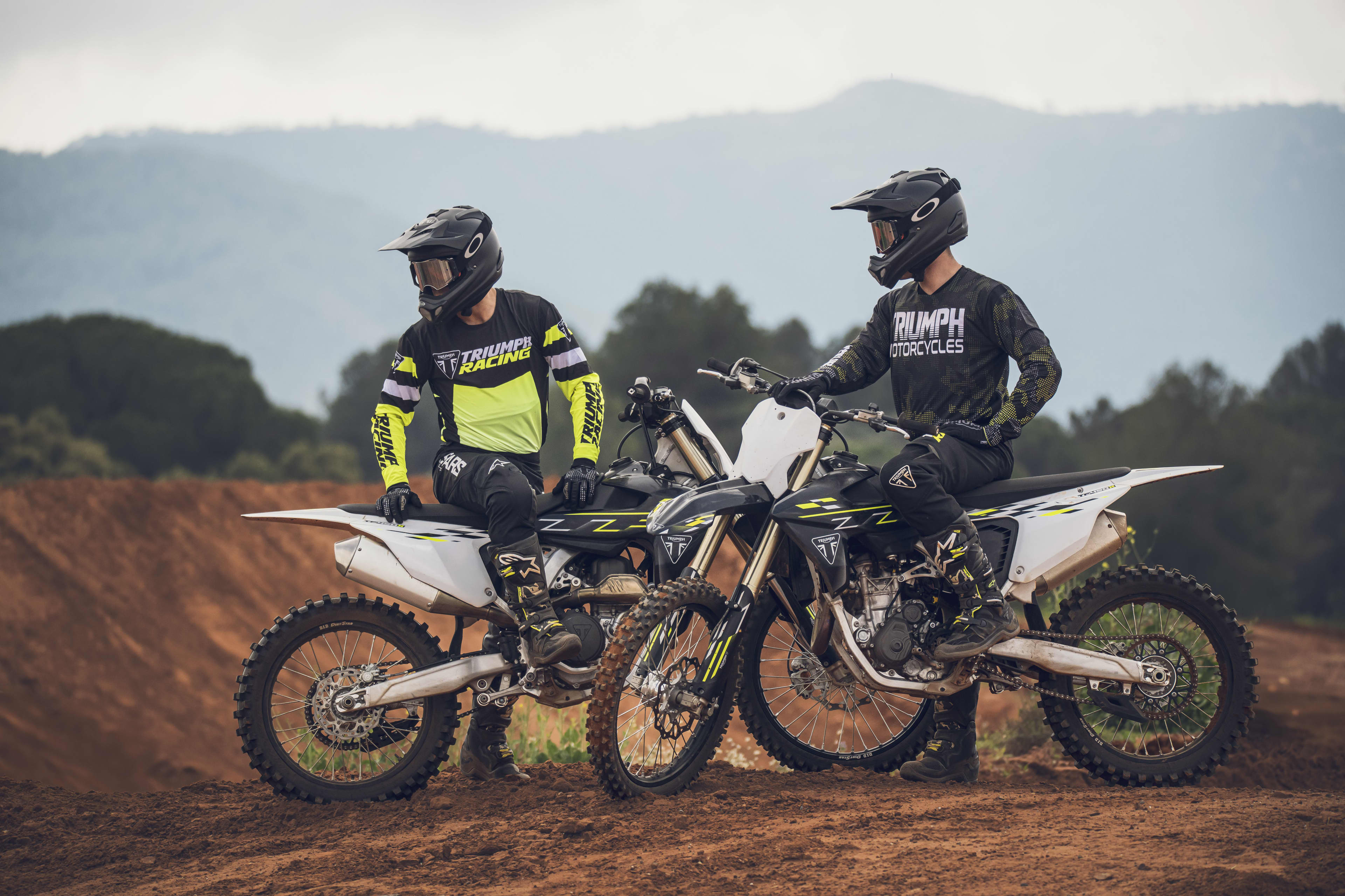 Motocross Two riders in full gear and helmets sitting on a Triumph TF 250-X and TF 450-X motocross bikes at an off-road dirt track with hills.