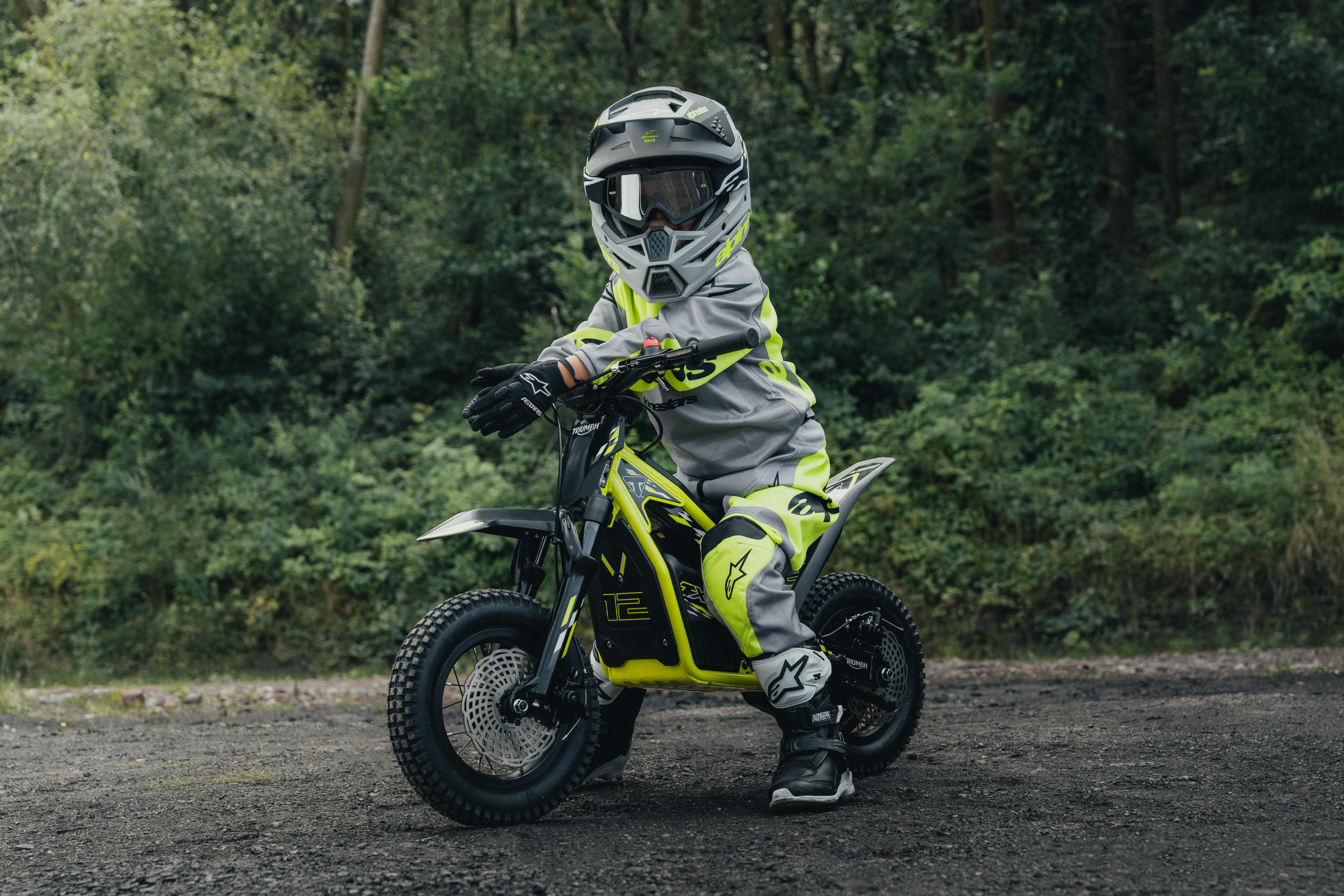 Start with confidence. Ung kører ved siden af en elektrisk Triumph TXP-12-offroadmotorcykel i en skov.
