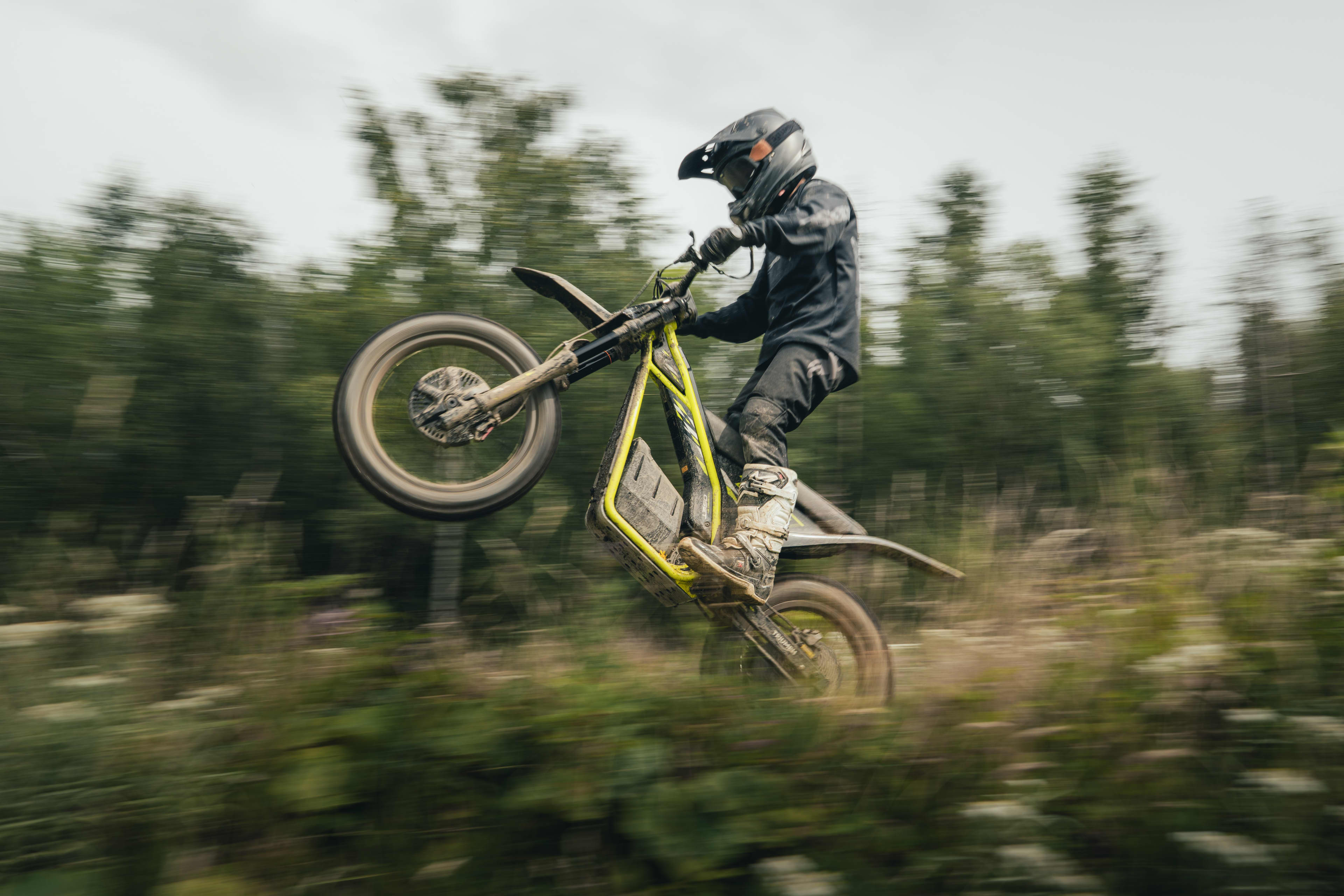 Ung kører laver en wheelie på en elektrisk Triumph TXP-20-offroadmotorcykel i en skov.