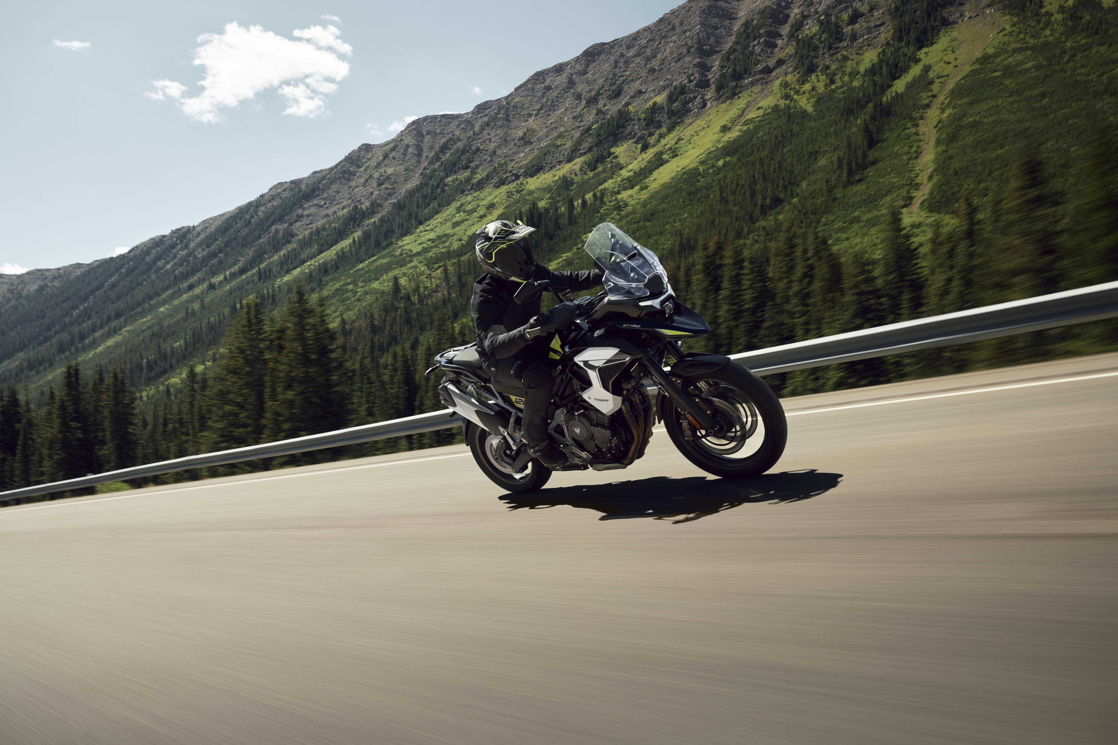 Side action shot of a rider on a Triumph Tiger 1200 GT Pro on a mountain highway with trees and mountains in the distance.