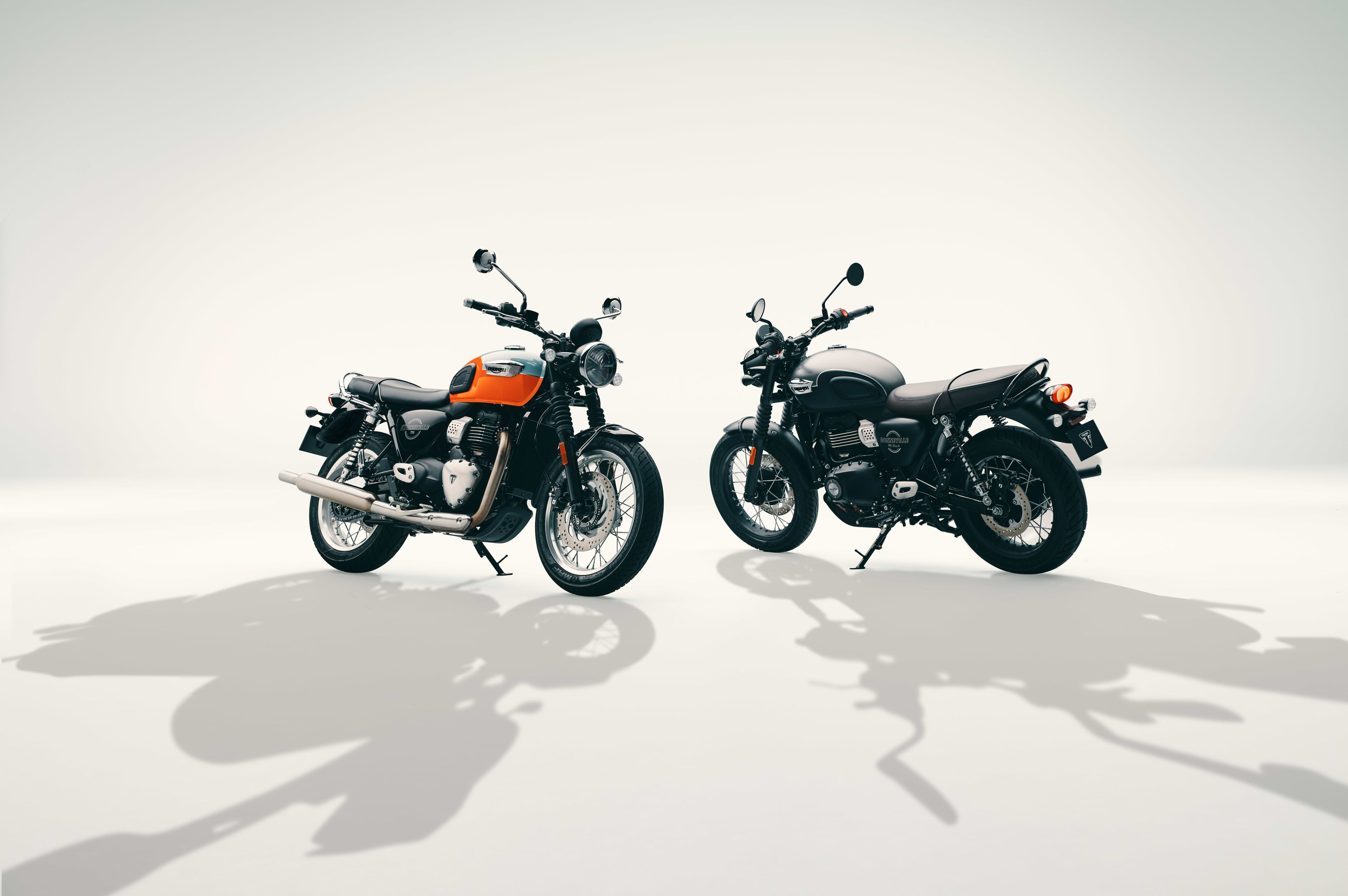 Studio shot of two Triumph Bonneville motorcycles—one orange and light blue T40, one matte black T40 Black—parked on a white background.