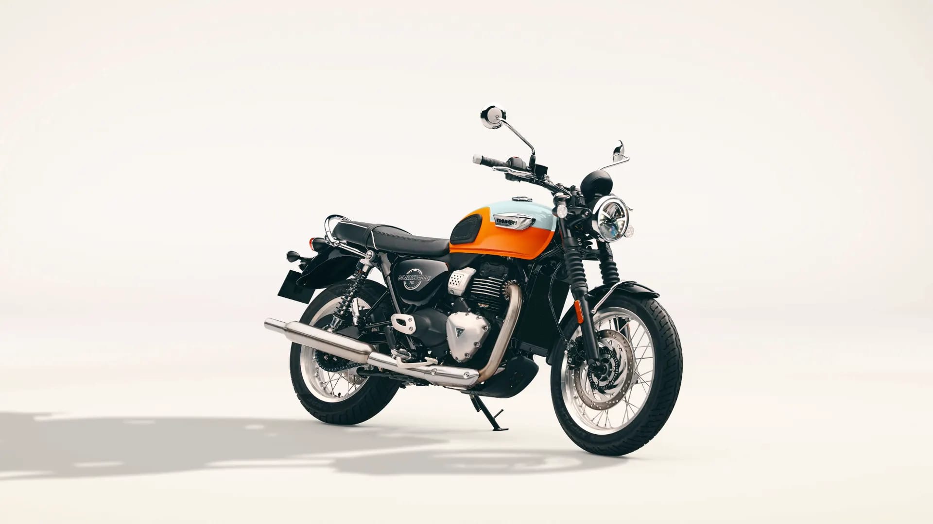 Studio shot of an orange and white Triumph Bonneville T40 motorcycle with chrome exhaust parked on a light background.