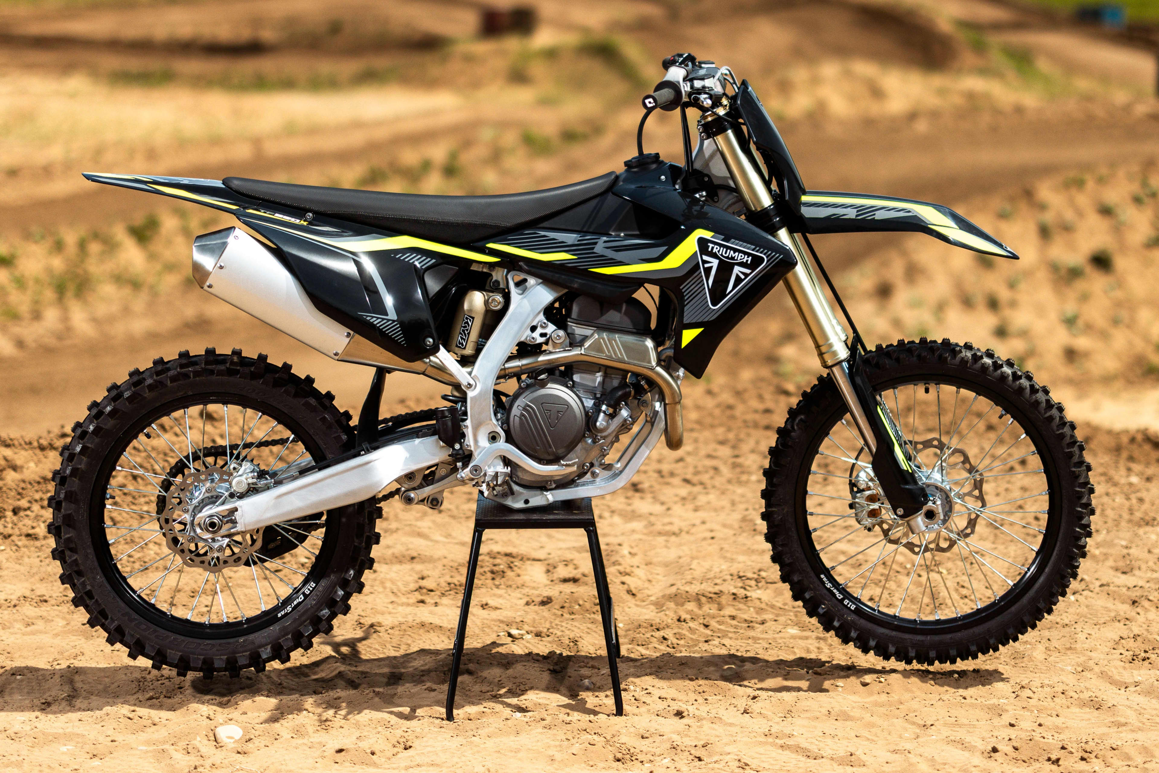 A side profile shot of a Triumph TF 250-X off-road bike resting on a central paddock stand at an outdoor dirt track.