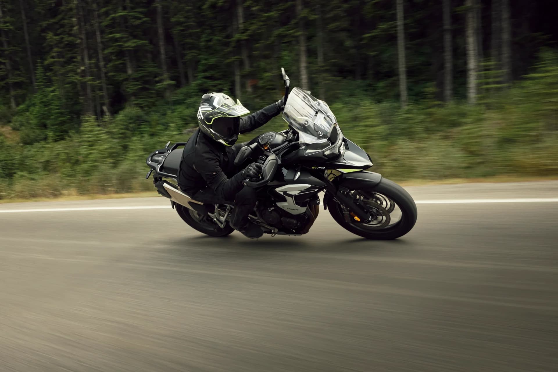 Dynamic action shot of a rider leaning a Triumph Tiger 1200 GT Pro through a turn along a forest-lined highway.