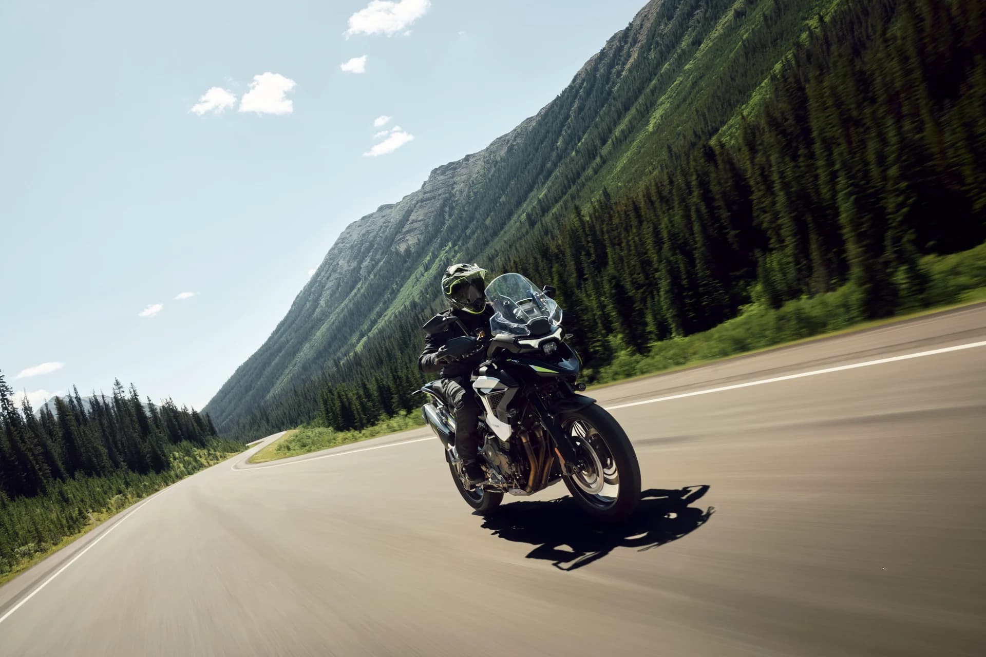 Wide-angle action shot showing a rider on a Triumph Tiger 1200 GT Pro cruising along a scenic mountain highway.