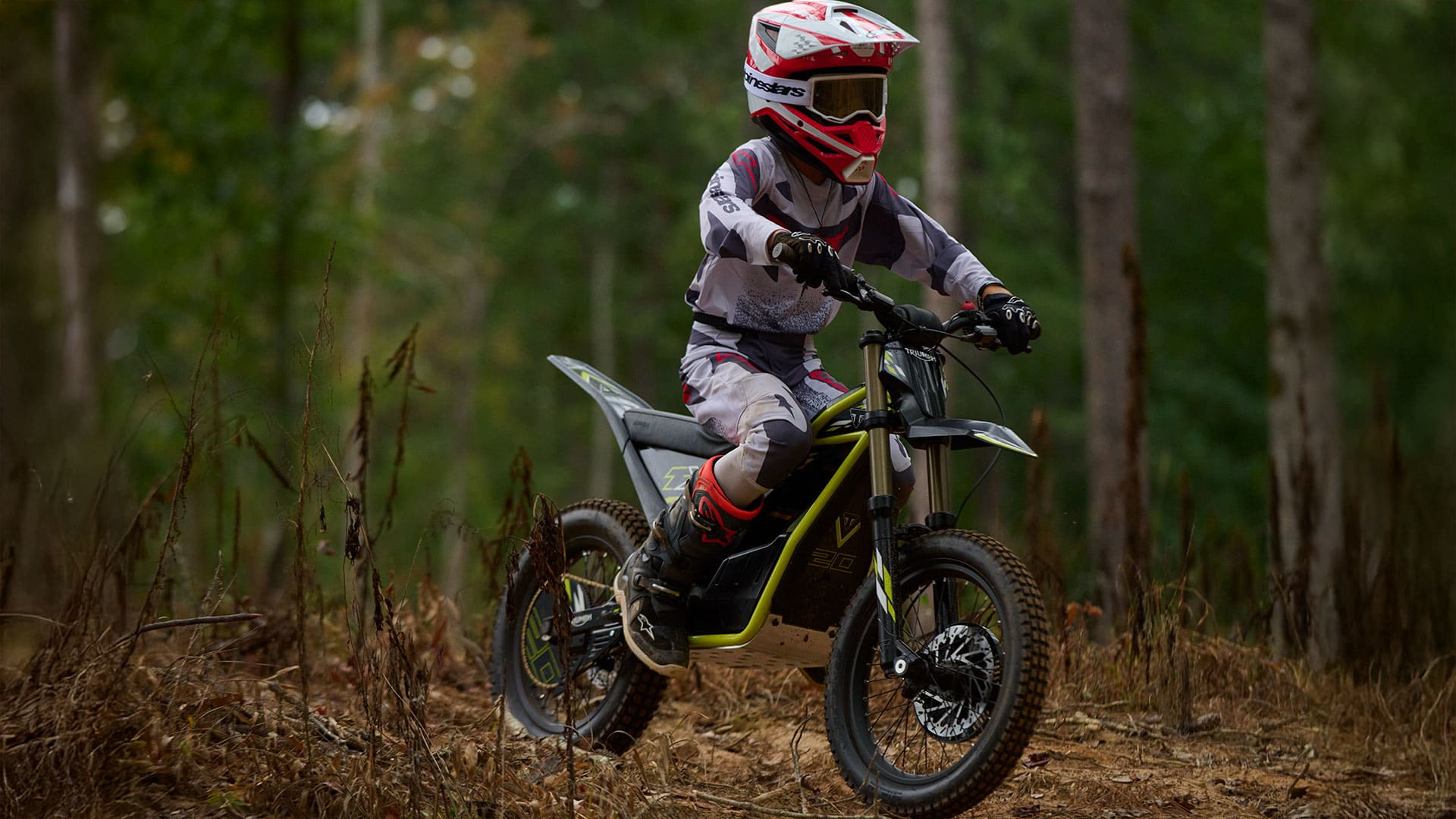Three-quarter action shot of a young rider fully geared in a red and white helmet, goggles, jersey, and boots riding a Triumph TXP-20 electric motorcycle along a woodland trail.