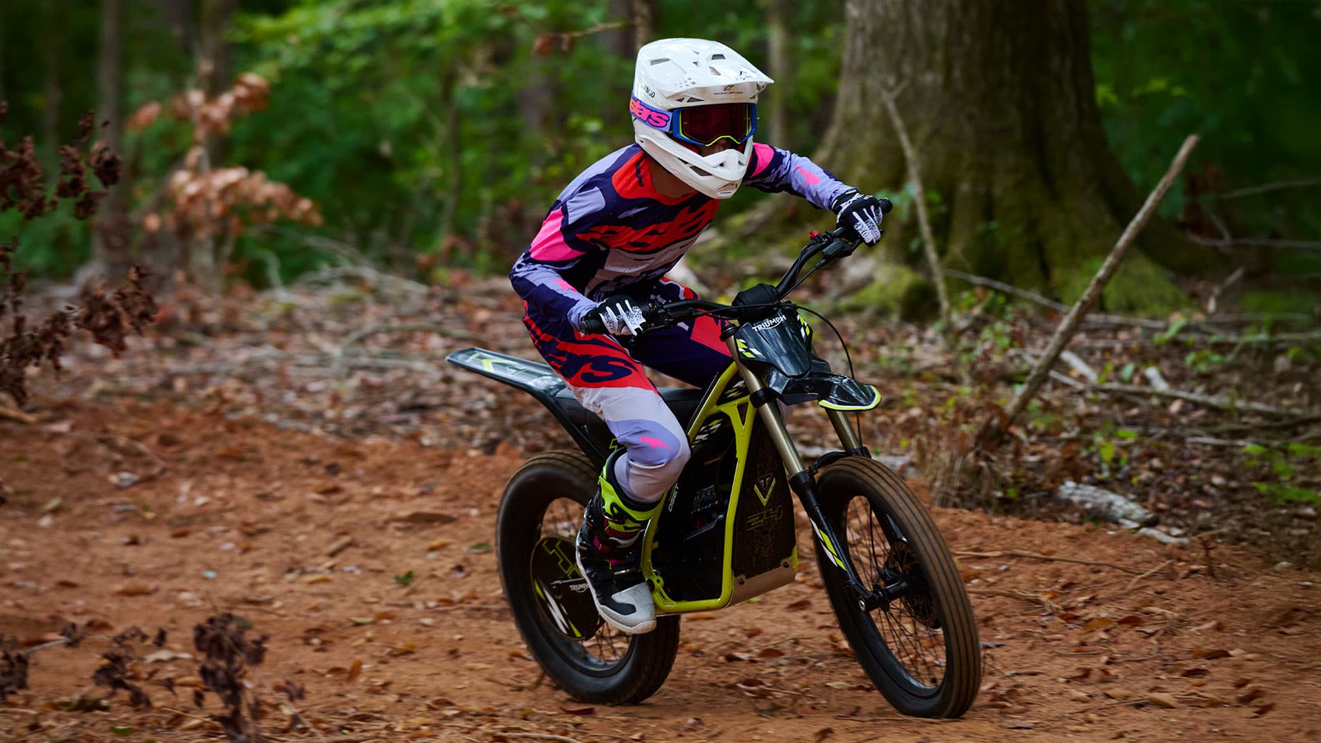 Big performance. Full-size capability. Three-quarter action shot of a young rider fully outfitted in a white helmet, goggles, and pink and purple gear riding a Triumph TXP-24 electric motorcycle on a dirt forest trail.
