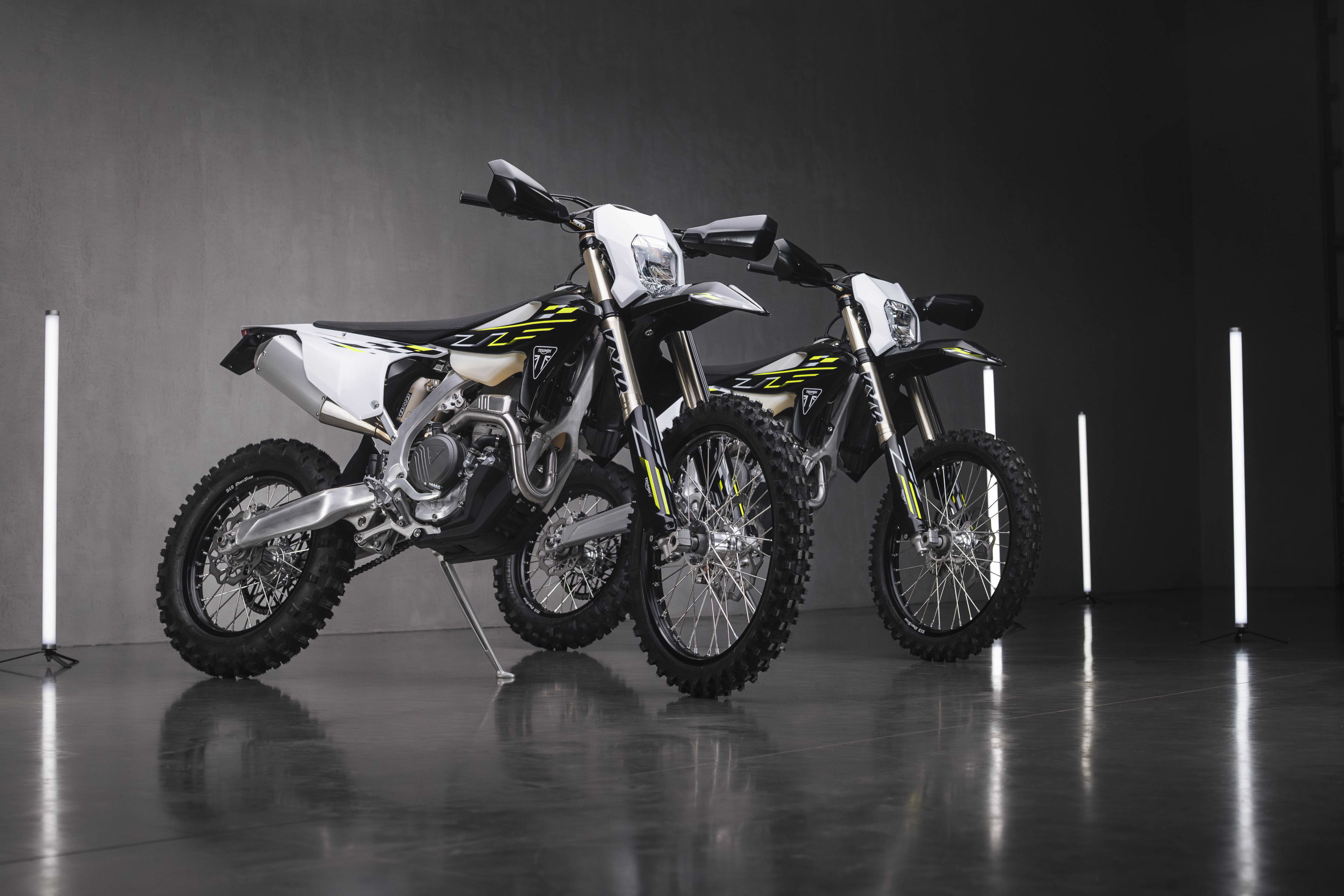 Two Triumph enduro motorbikes parked on a glossy dark floor in a studio setting with vertical tube lights.