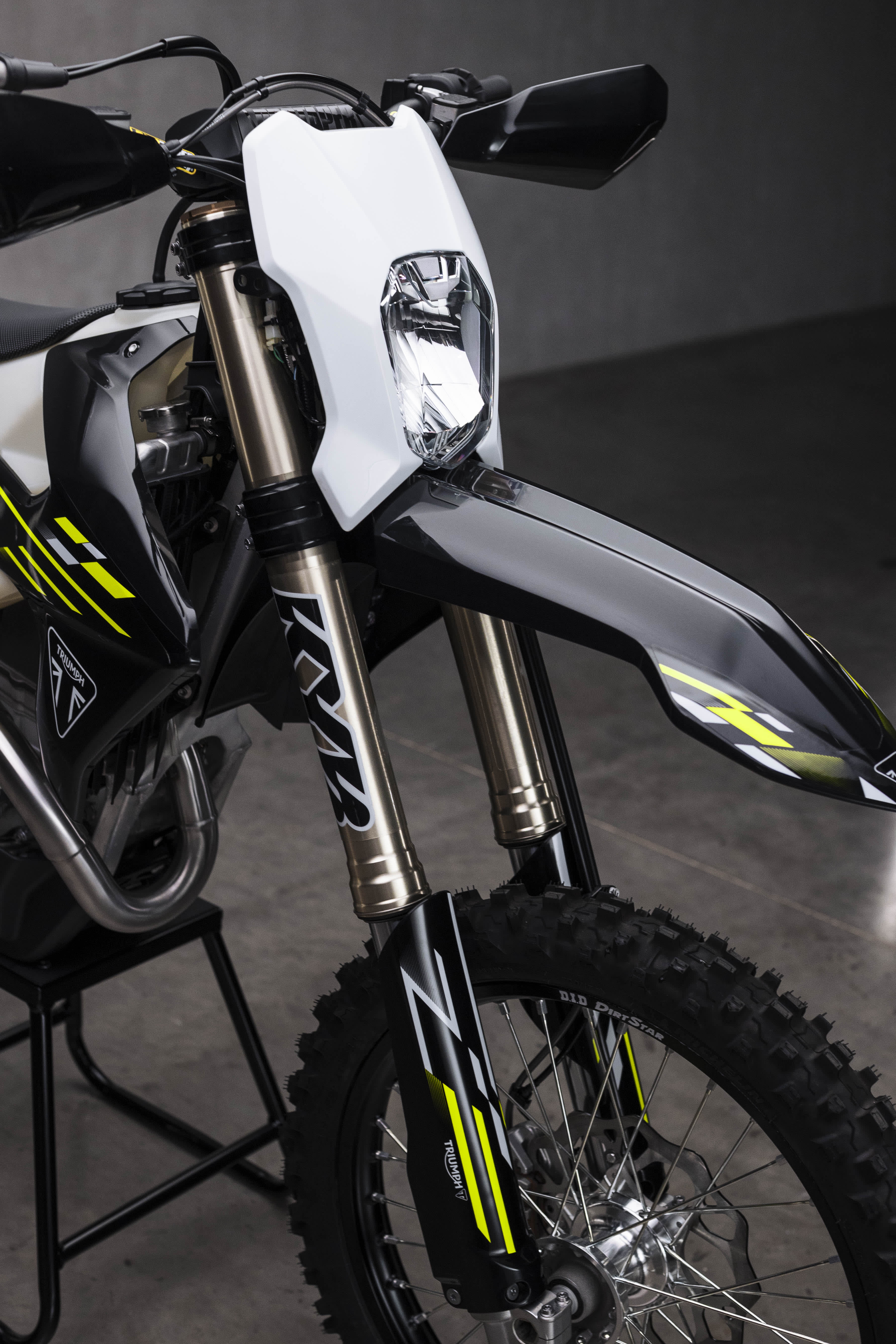 A close-up shot of the front assembly of a Triumph TF 450-E enduro motorcycle, featuring its white headlight mask, high-mounted black front fender, and golden front forks with KYB suspension decals.