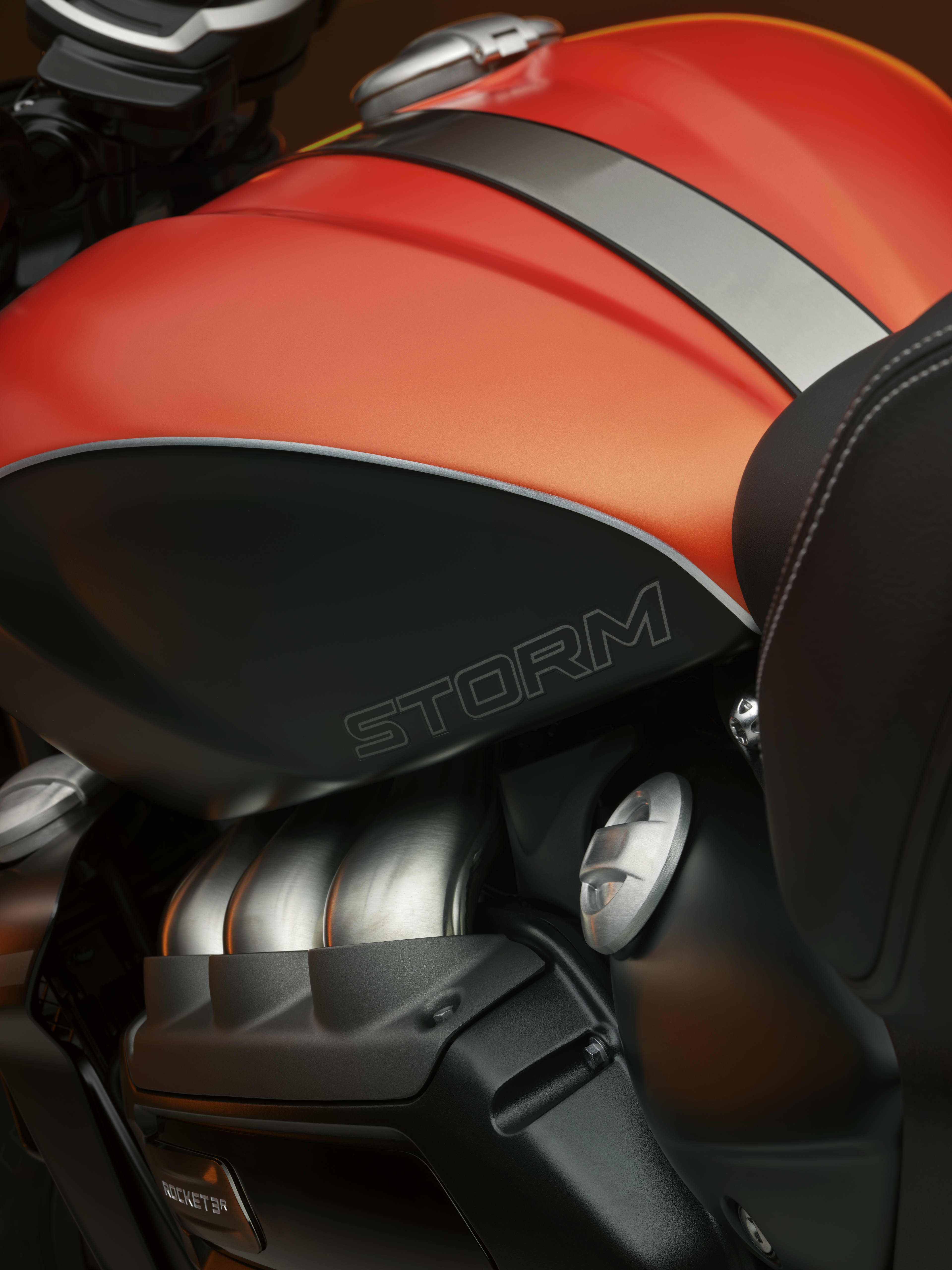 A close-up, artistic shot of a Triumph Rocket 3 Storm R motorcycle, highlighting its orange and black fuel tank with the "Storm" logo alongside a view of its massive blacked-out engine components.