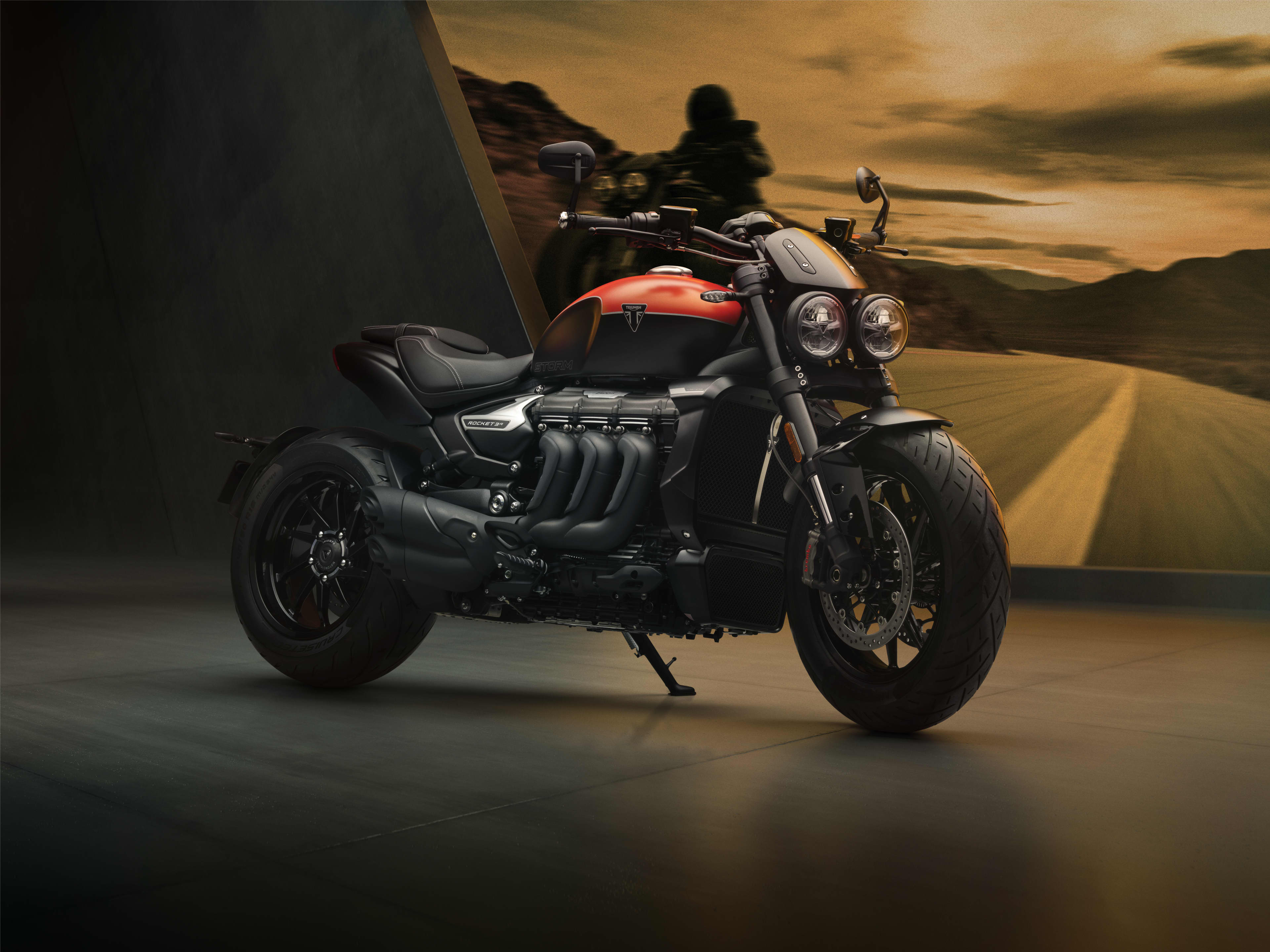 A black and red Triumph Rocket 3 Storm R cruiser motorcycle parked on a showroom floor in front of a scenic desert highway backdrop.