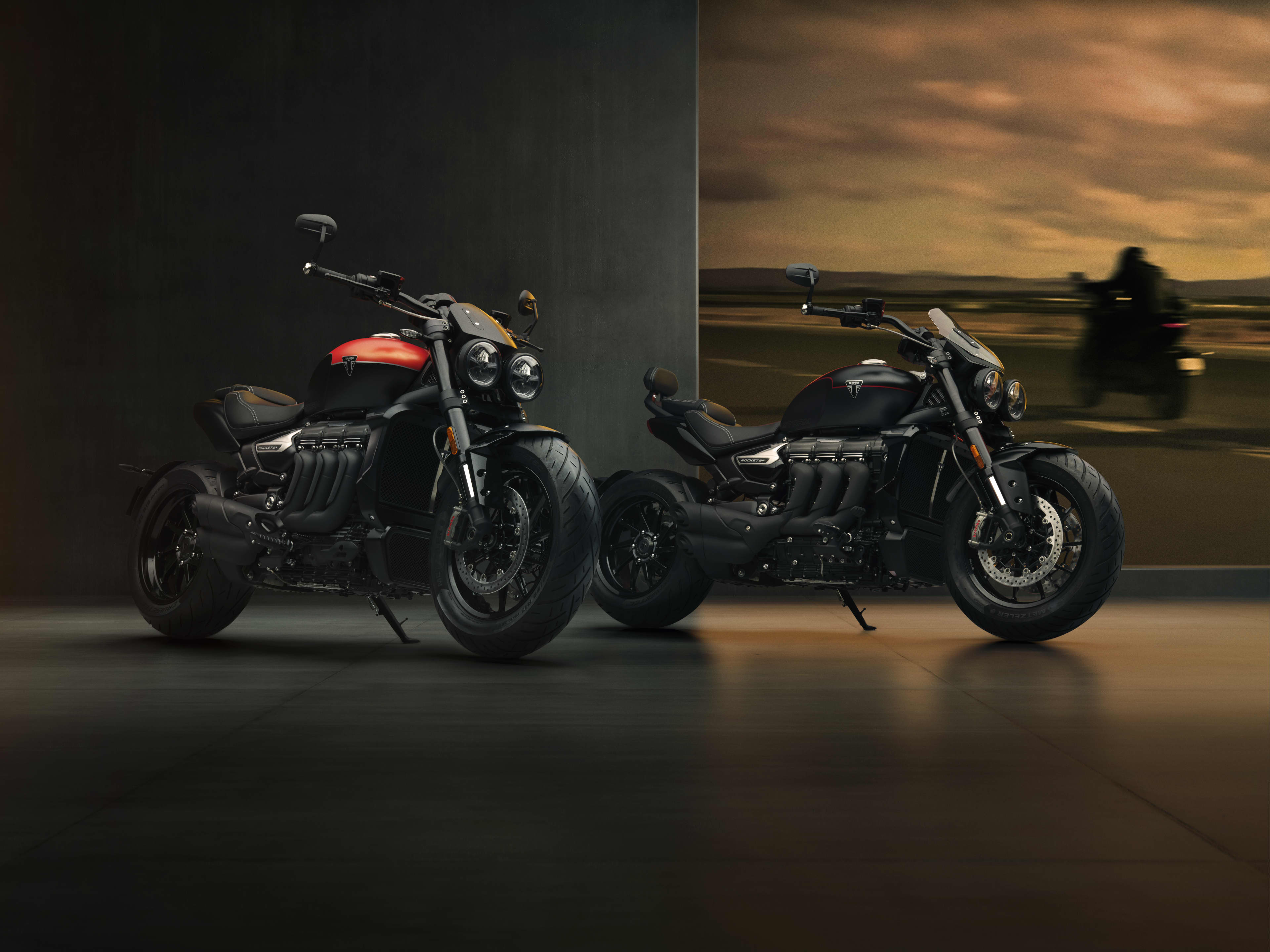 Two Triumph Rocket 3 Storm motorcycles—the R model with a red tank accent and the GT model in all-black—parked side-by-side in a dimly lit studio showroom.