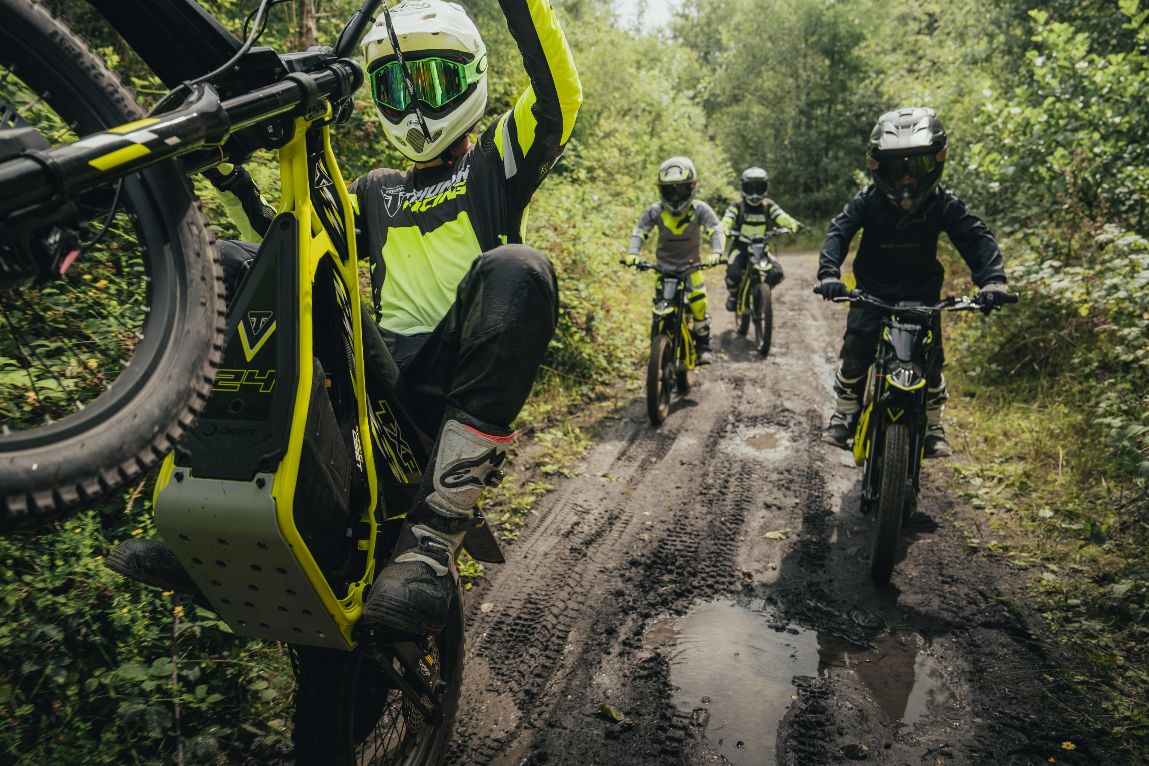 A group of young riders navigating a muddy, tree-lined trail on neon yellow Triumph TXP electric dirt bikes, with the lead rider performing a wheelie.