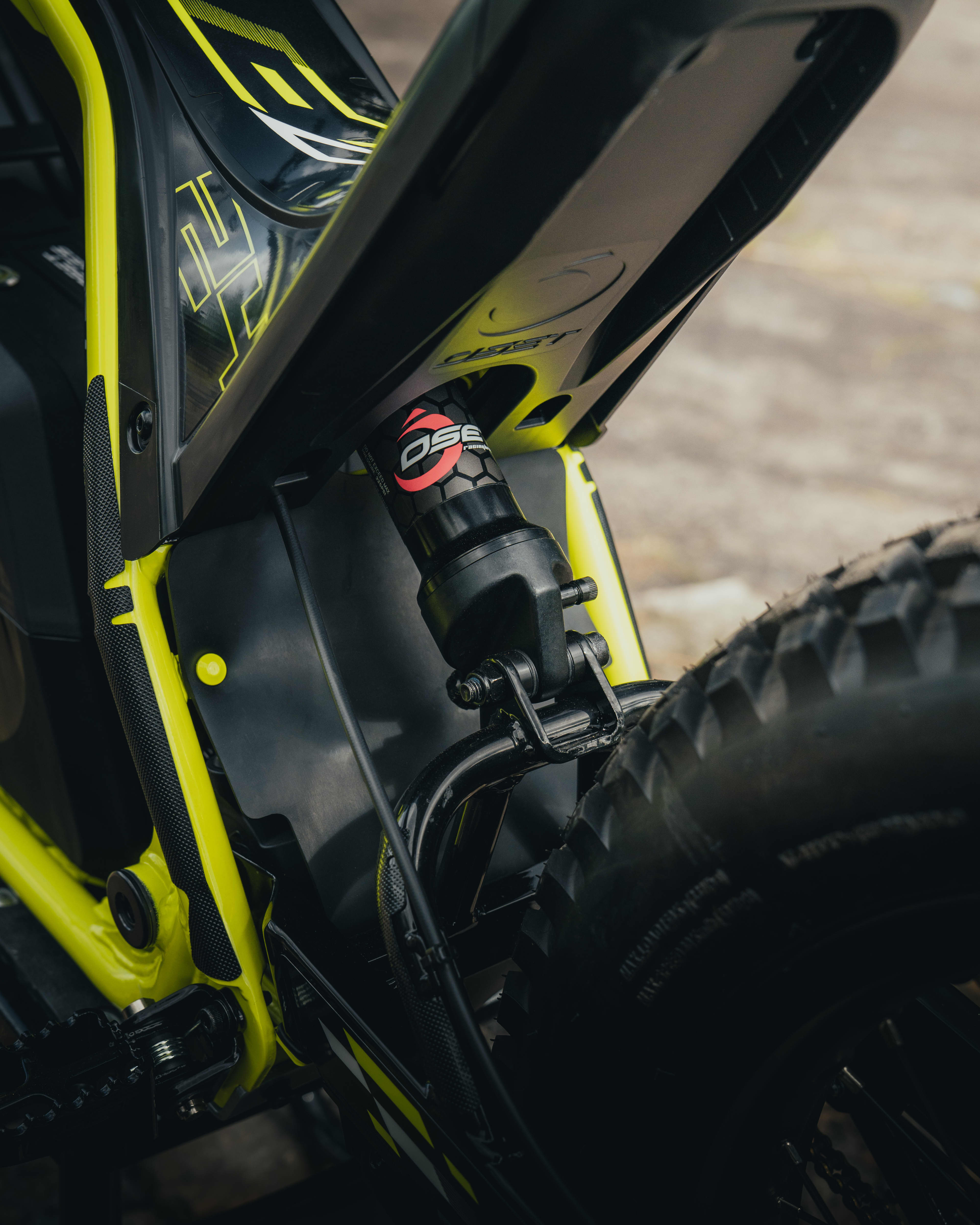 A close-up shot of the rear suspension setup on a Triumph TXP-24 electric bike, showcasing the DNM rear shock absorber nestled within the neon yellow frame near the knobby rear tire.