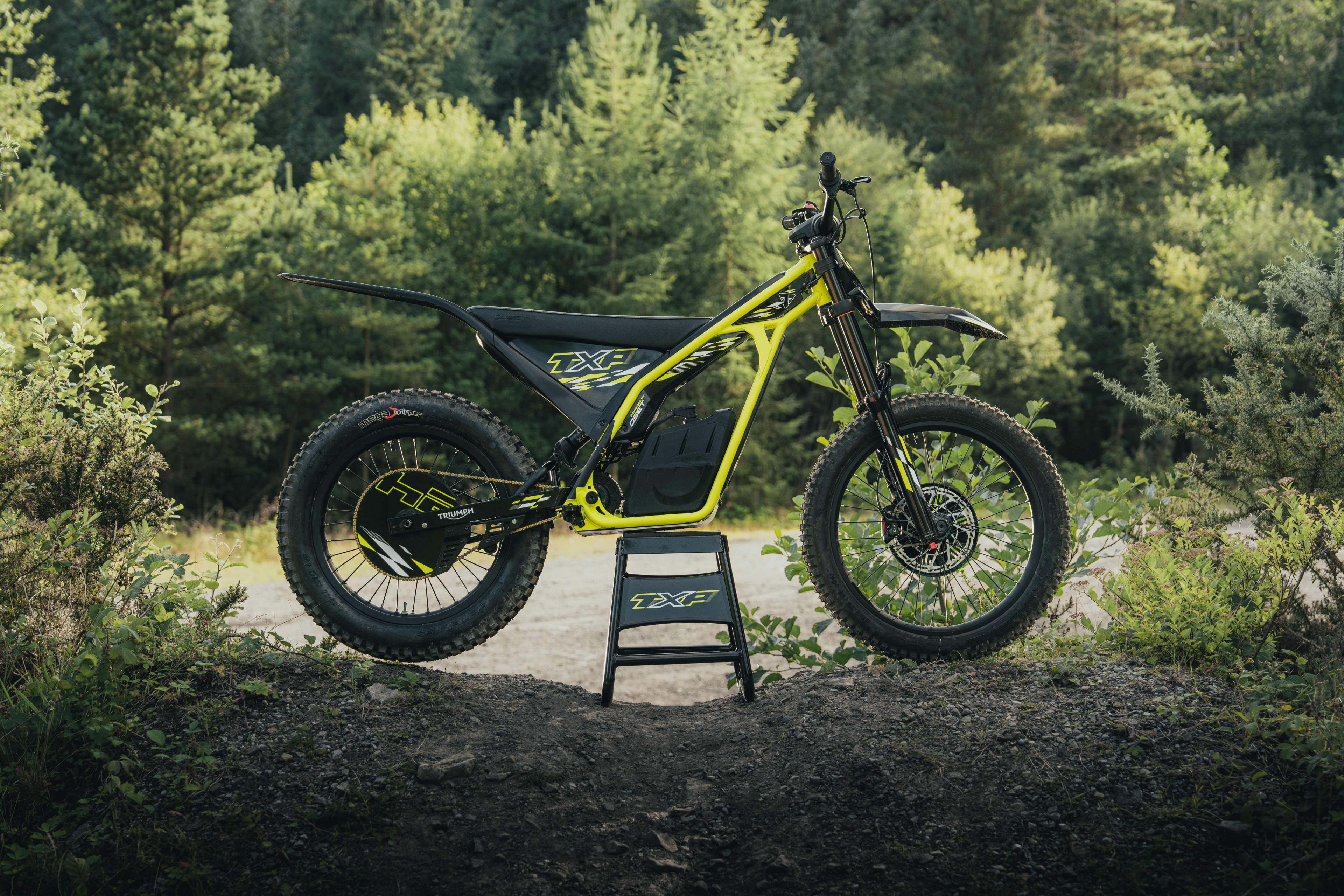 A side profile shot of a neon yellow and black Triumph TXP electric off-road bike resting on a paddock stand on a dirt path with a lush forest background.