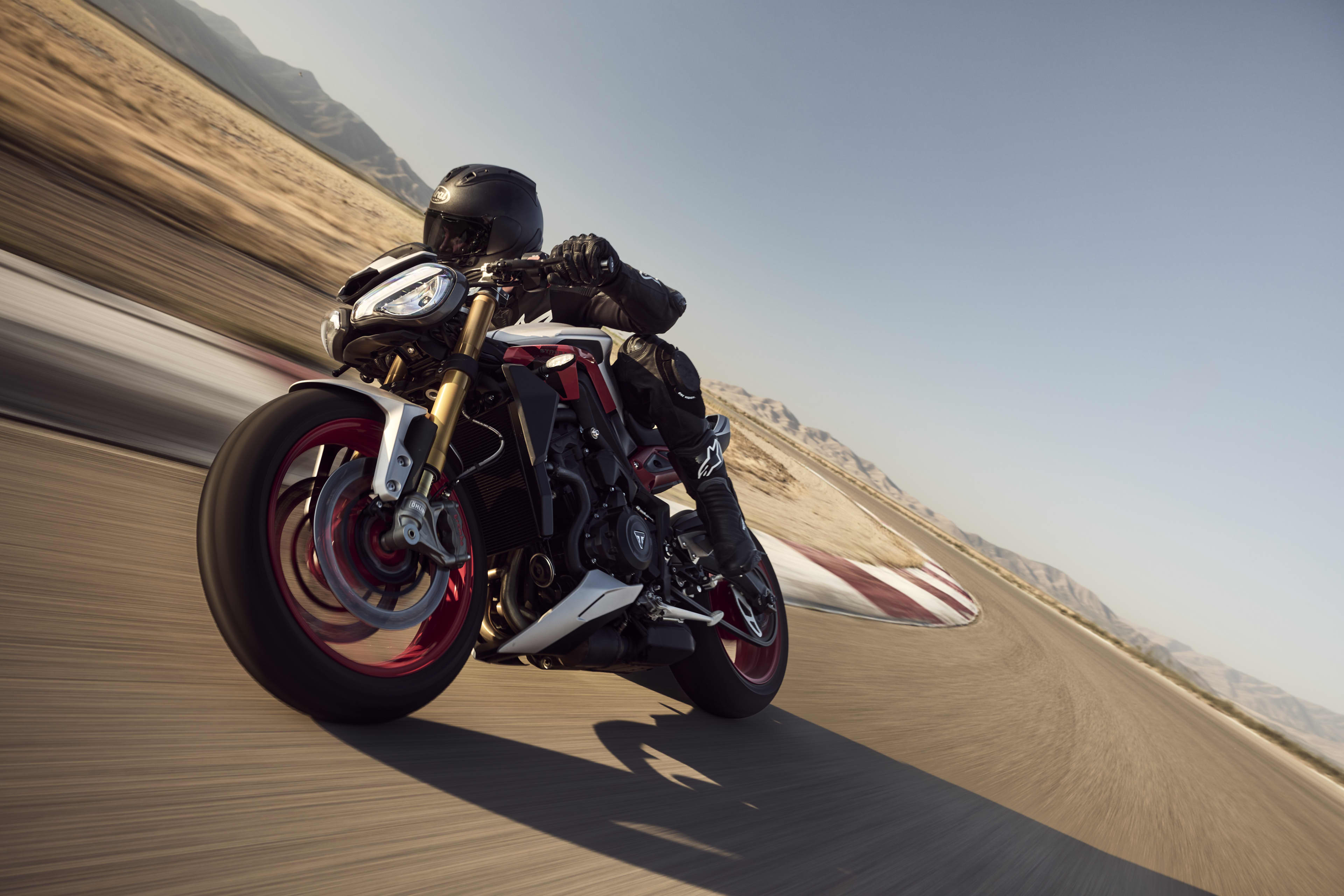 A motorcyclist in black leathers leaning heavily into a high-speed turn on a Triumph Street Triple 765 RX sportbike, showcasing its distinctive red wheels.
