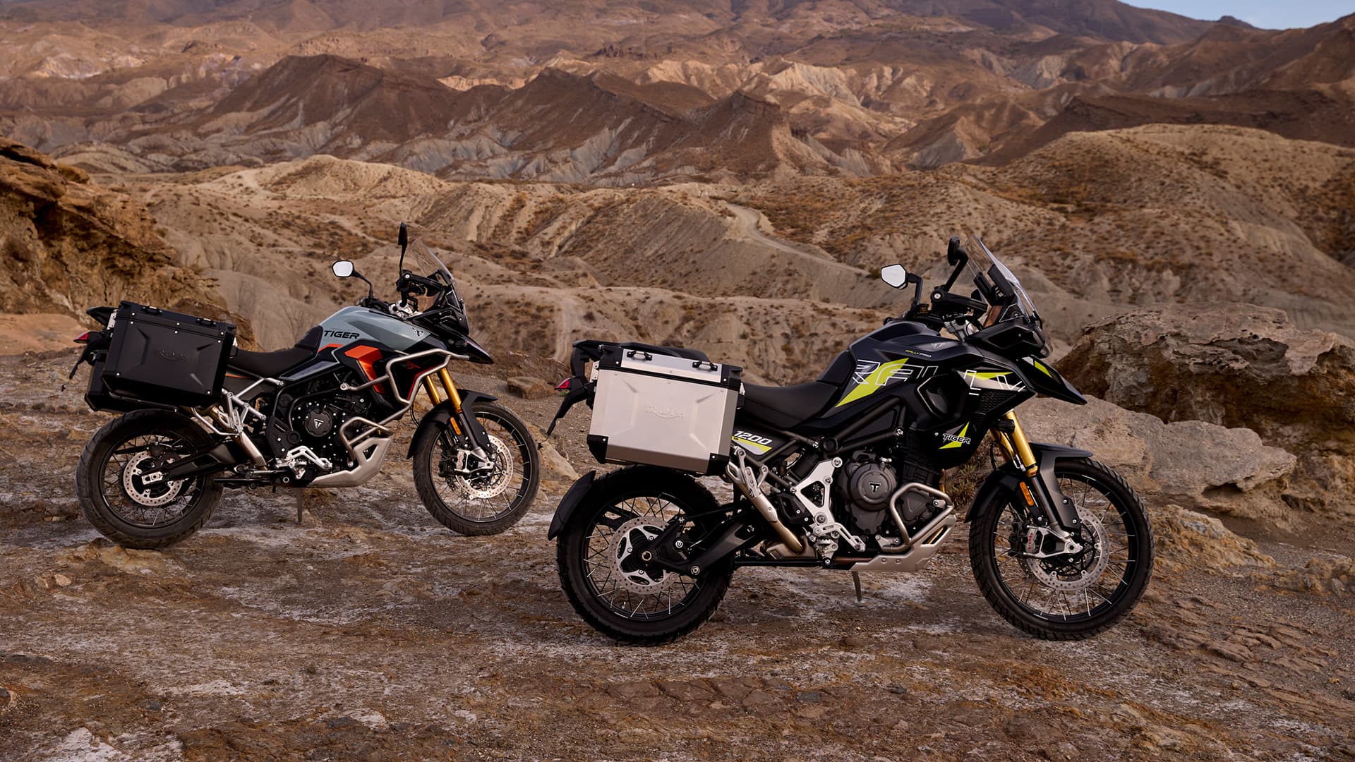 Two Triumph adventure motorcycles—a Tiger 900 and a Tiger 1200—parked on a rocky desert landscape at sunset with mountains in the background.