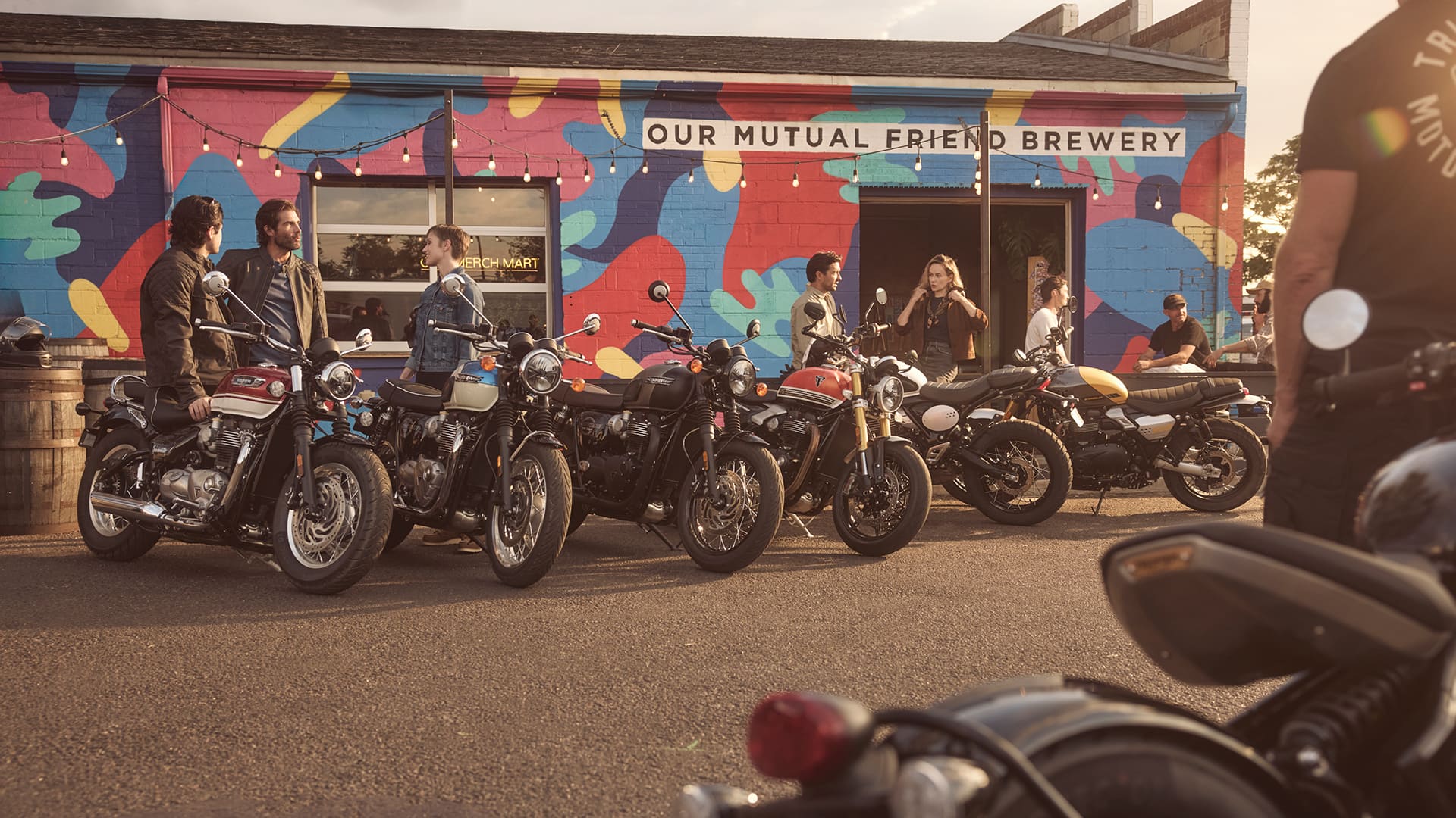 A line of classic Triumph motorcycles parked in front of Our Mutual Friend Brewery, with people socializing in the background under an outdoor mural.