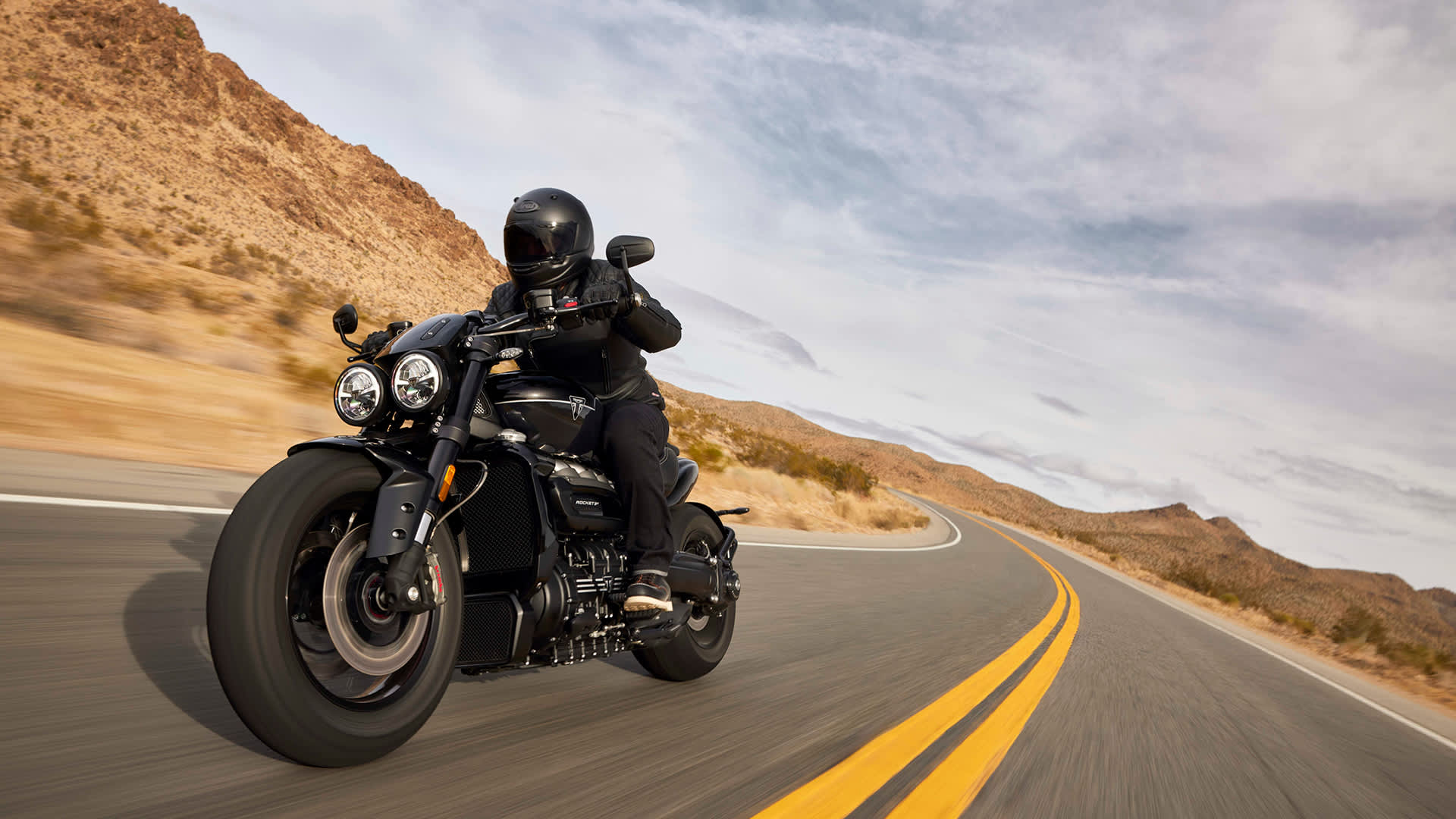 A motorcyclist wearing a black helmet and gear rides a Triumph Rocket 3 Storm GT along a curved desert highway with mountains in the background.