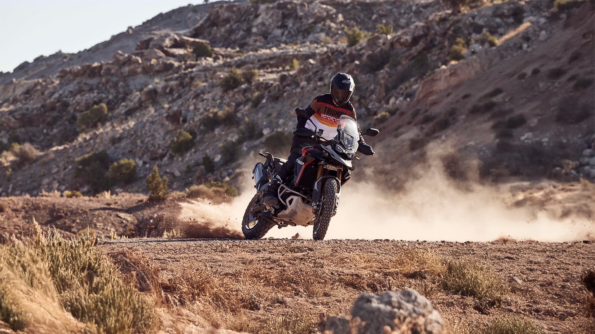 An adventure motorcyclist kicks up dust while riding a Triumph Tiger 900 Rally Pro on an off-road, rugged desert trail.