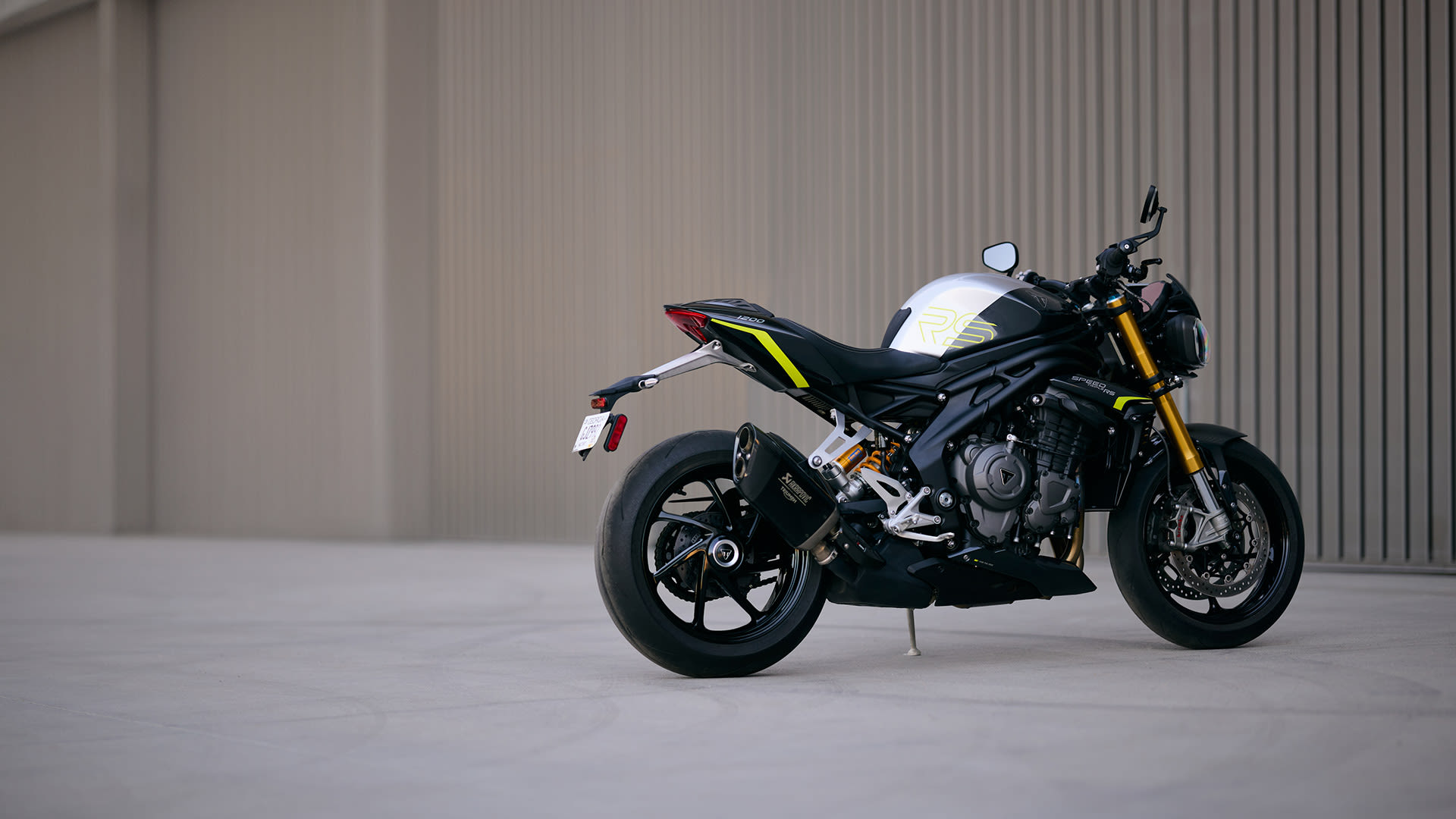 Side profile of a black and silver Triumph Speed Triple RS motorcycle with yellow accents parked outdoors.