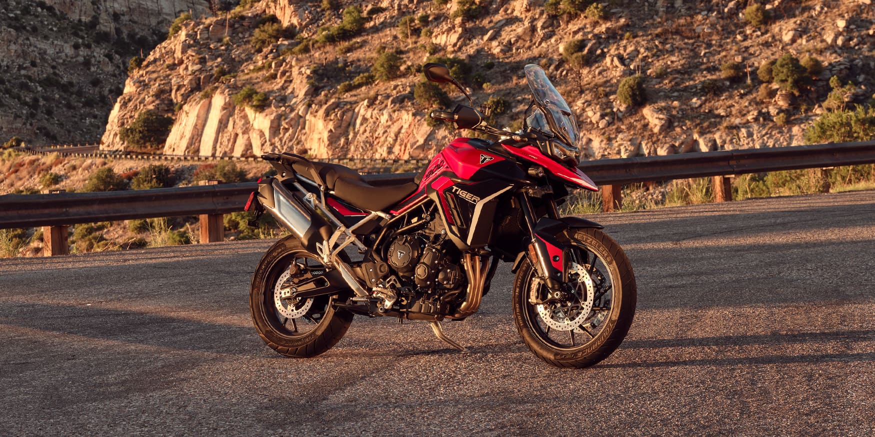 A red and black Triumph Tiger 900 GT adventure motorcycle parked on an asphalt road with rocky cliffs in the background.