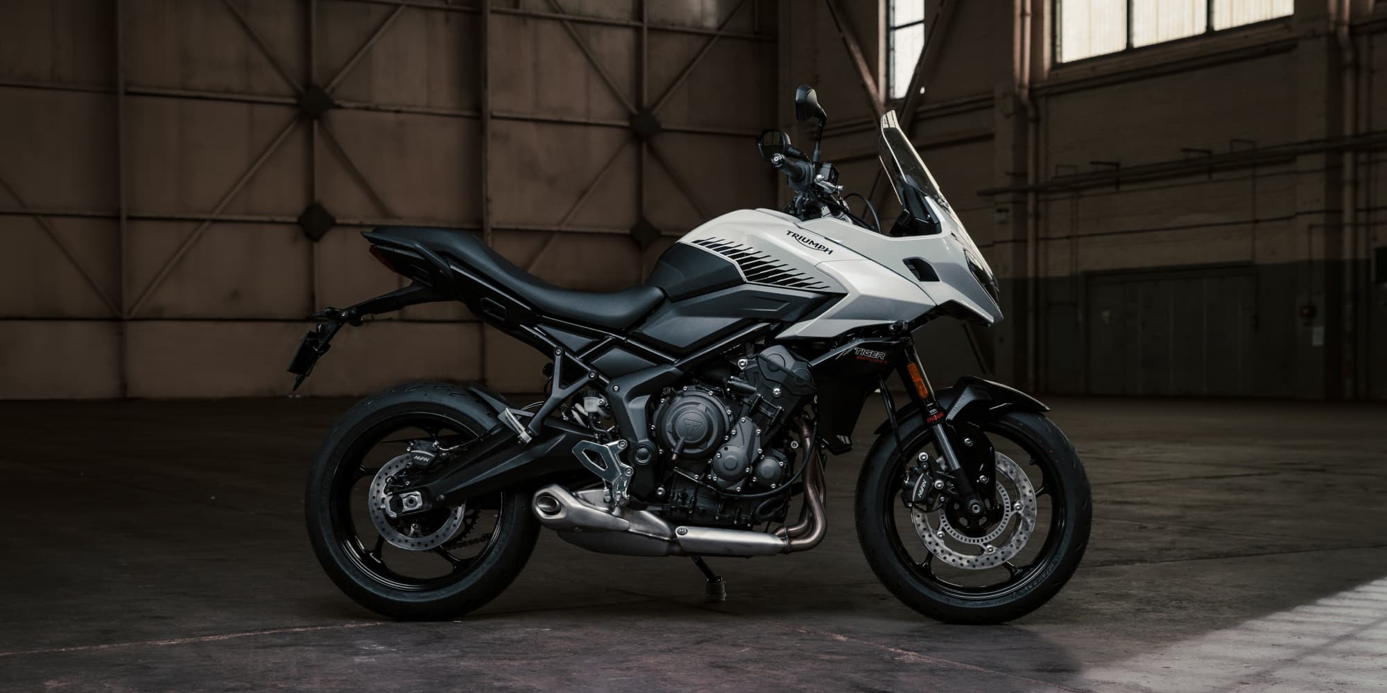 Side profile of a white and black Triumph Tiger Sport 660 motorcycle parked in an industrial hangar.