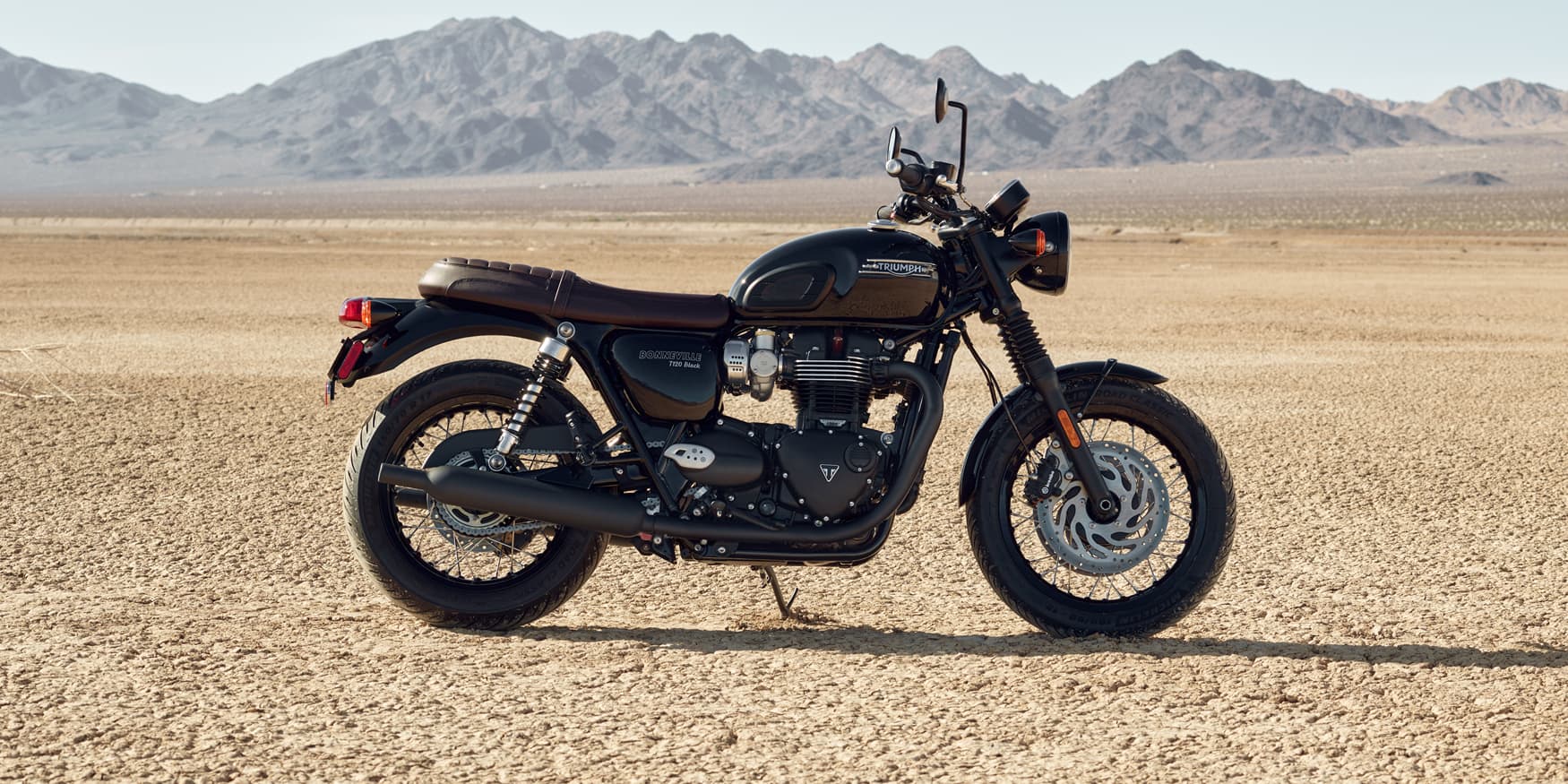 Side profile of a black Triumph Bonneville T120 motorcycle with a brown leather seat parked in a desert landscape.
