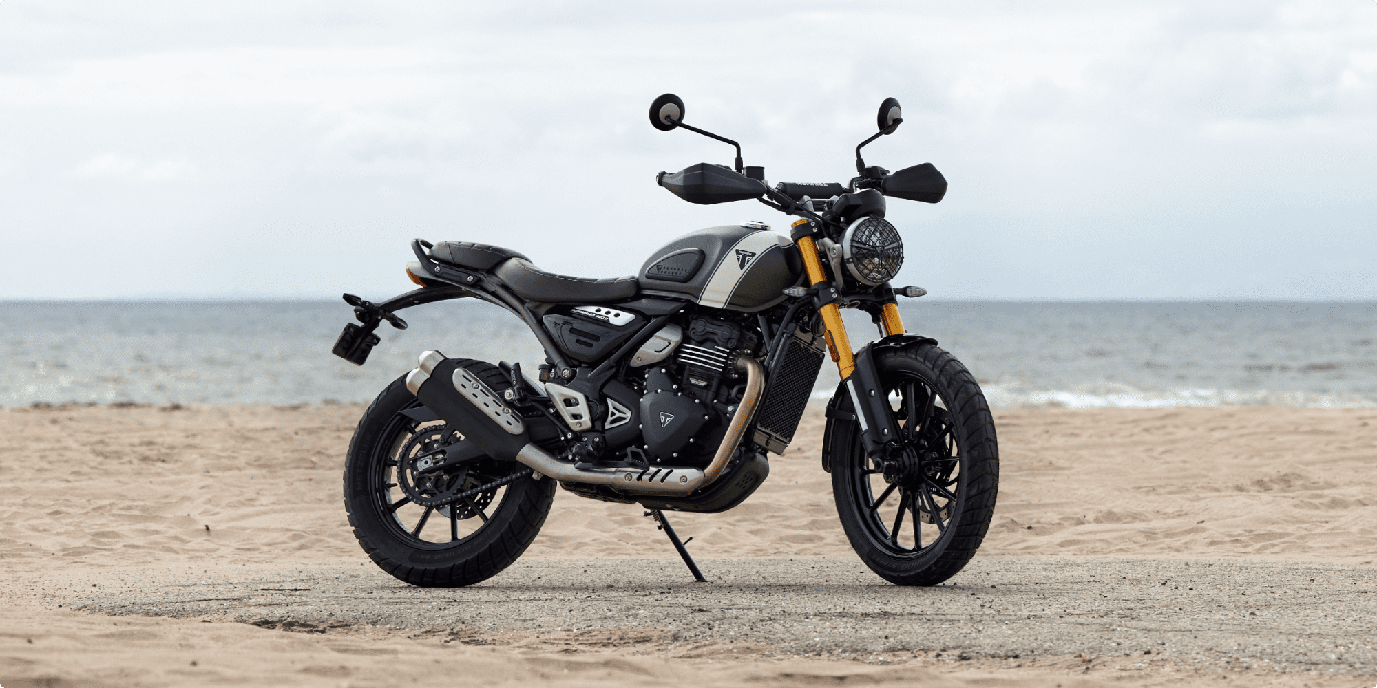 Green Triumph Scrambler 400 X motorcycle with gold front forks parked on the beach near to the ocean.