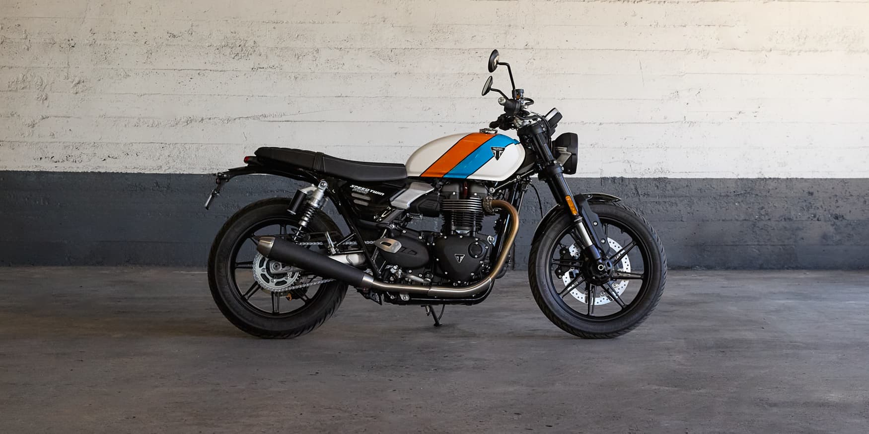 Profile view of a custom Triumph Speed 400 motorcycle featuring orange and blue fuel tank stripes in a garage.
